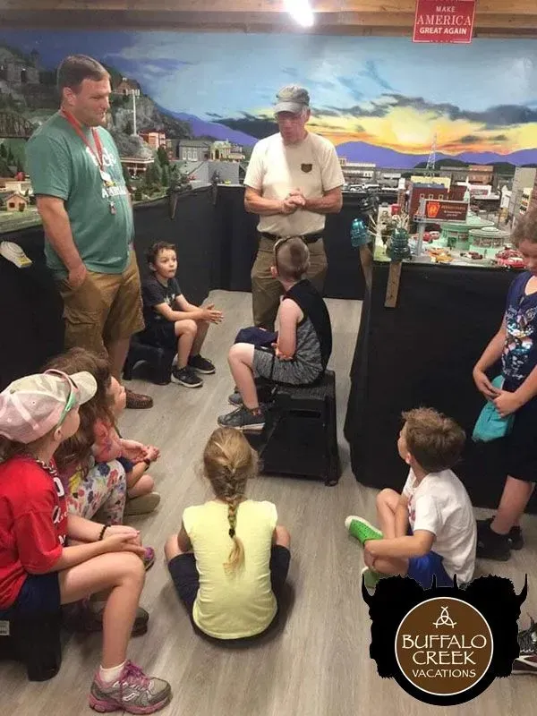 Children and two adults at a model train display, gathered for a demonstration.