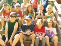 Group of children smiling, posing on wooden stairs, wearing casual clothes, and sunglasses.