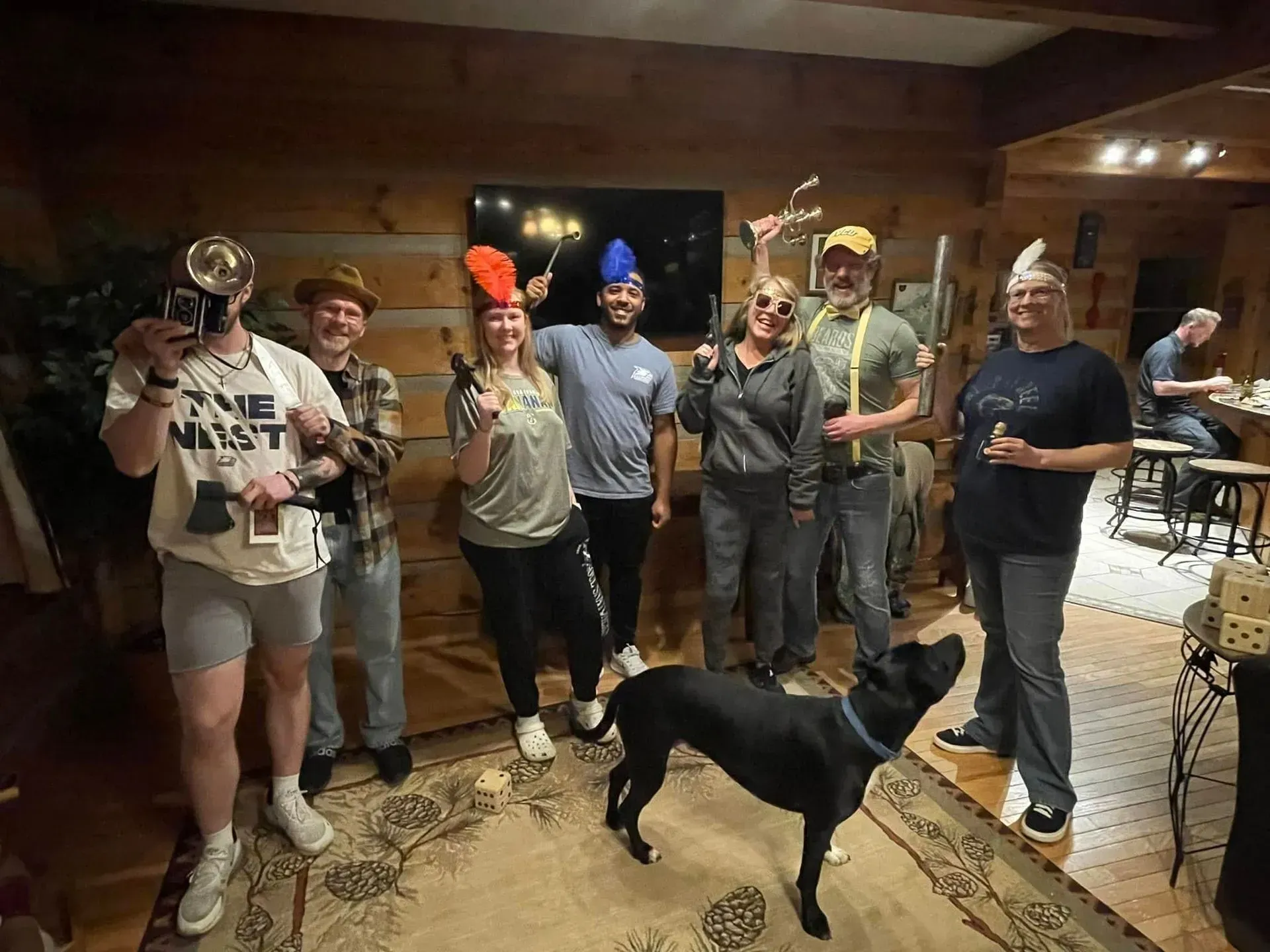 Group of friends in a wood-paneled room wearing novelty hats, posing with a dog.