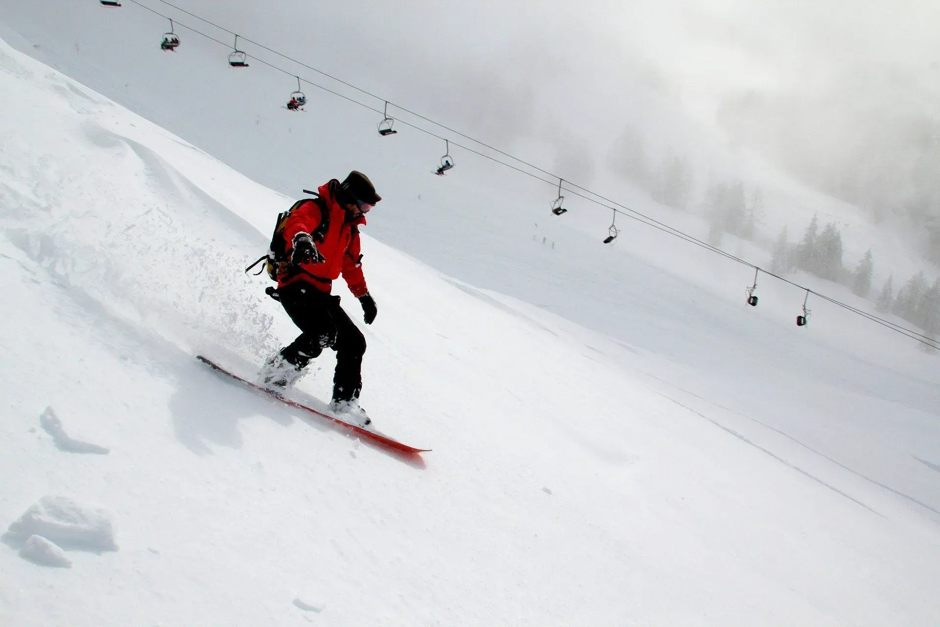 Snowboarder in red jacket carves down a snowy mountain slope with ski lift overhead.
