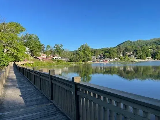Wooden walkway alongside a calm lake reflecting the sky and distant buildings and mountains under a blue sky.