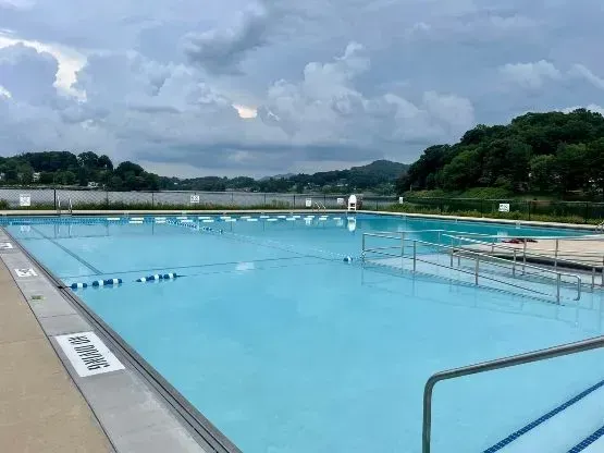 Empty outdoor swimming pool on a cloudy day, with a lake and trees in the background.