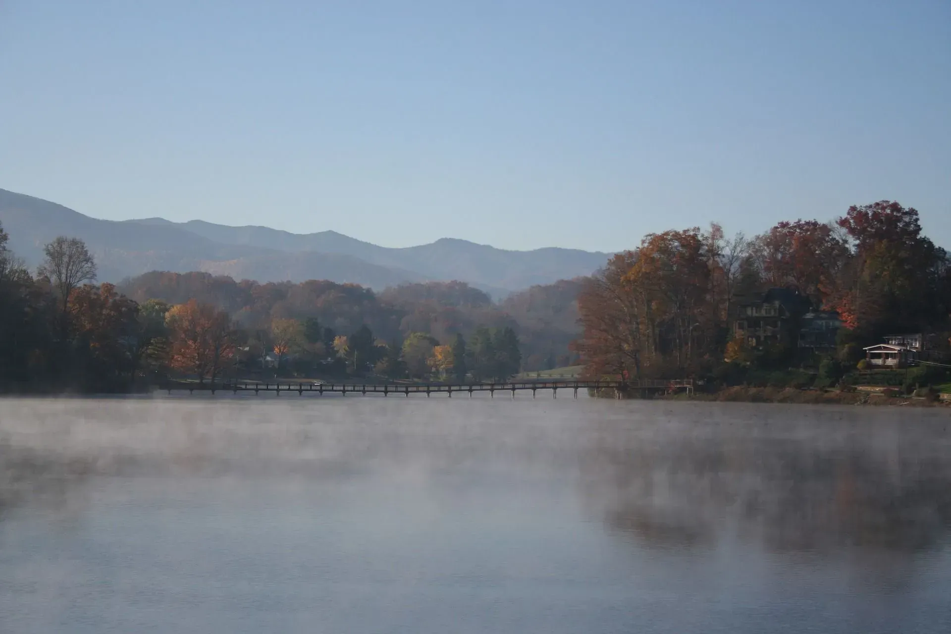 Foggy lake with a bridge, trees, and distant mountains under a clear blue sky.