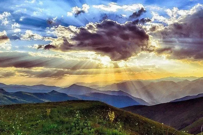 Sunlight streams through clouds over mountain ranges and a grassy foreground.