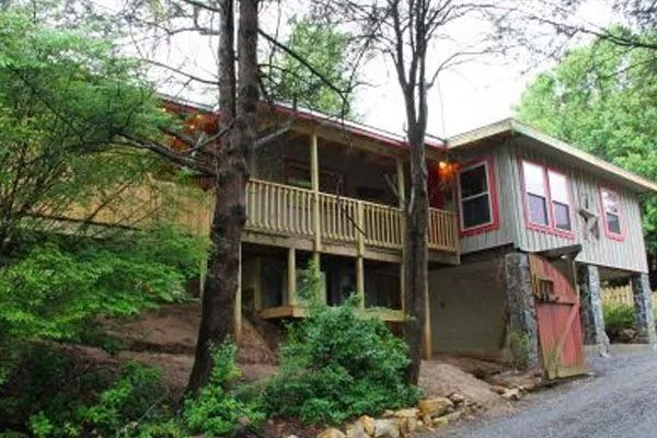 Cabin with wooden porch, surrounded by trees, set on a hillside. Red door and window trim.