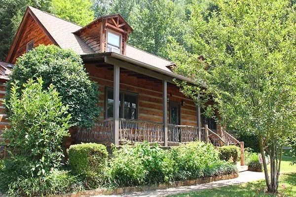 Log cabin nestled in lush greenery, featuring a porch and small dormer window.