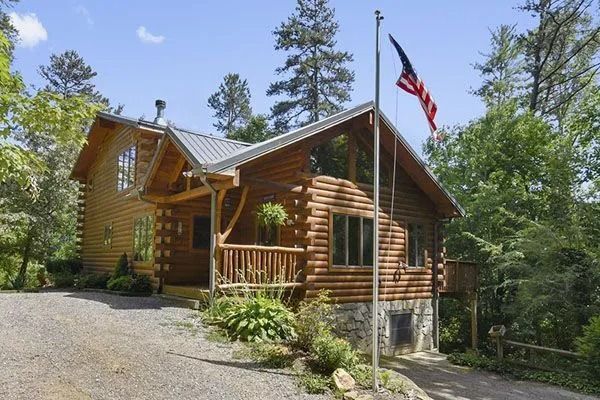 Log cabin with American flag, gravel driveway, surrounded by trees under a sunny sky.