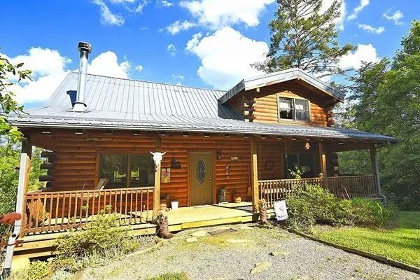 Log cabin with wraparound porch, metal roof, surrounded by trees, under a blue sky with clouds.