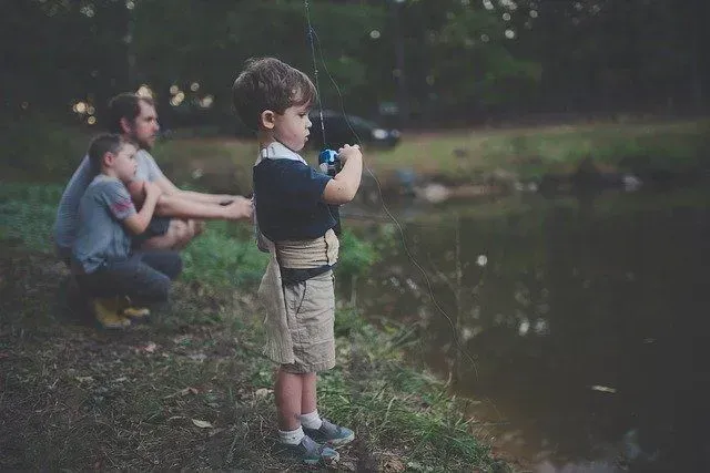 A young boy fishing, guided by a man, with another boy watching. Overcast day at a pond.