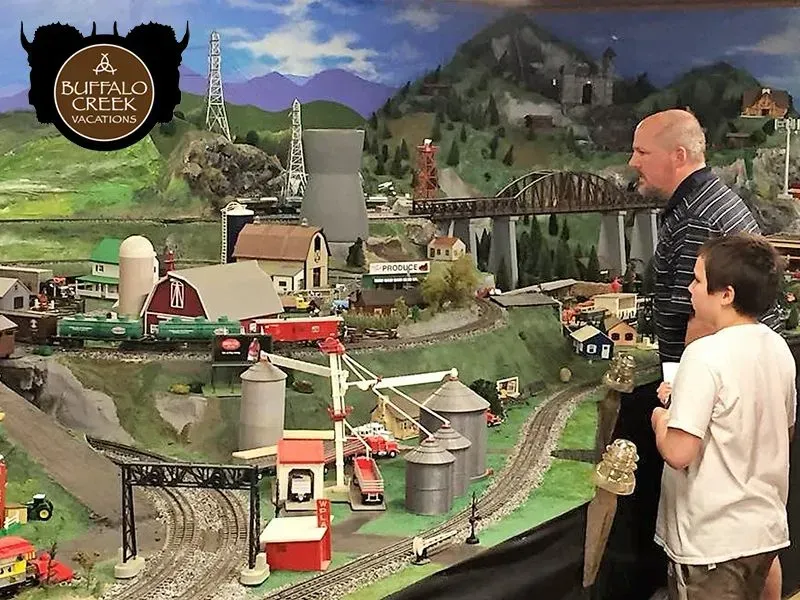 A man and a boy looking at a model train set with buildings, a bridge, and mountains.