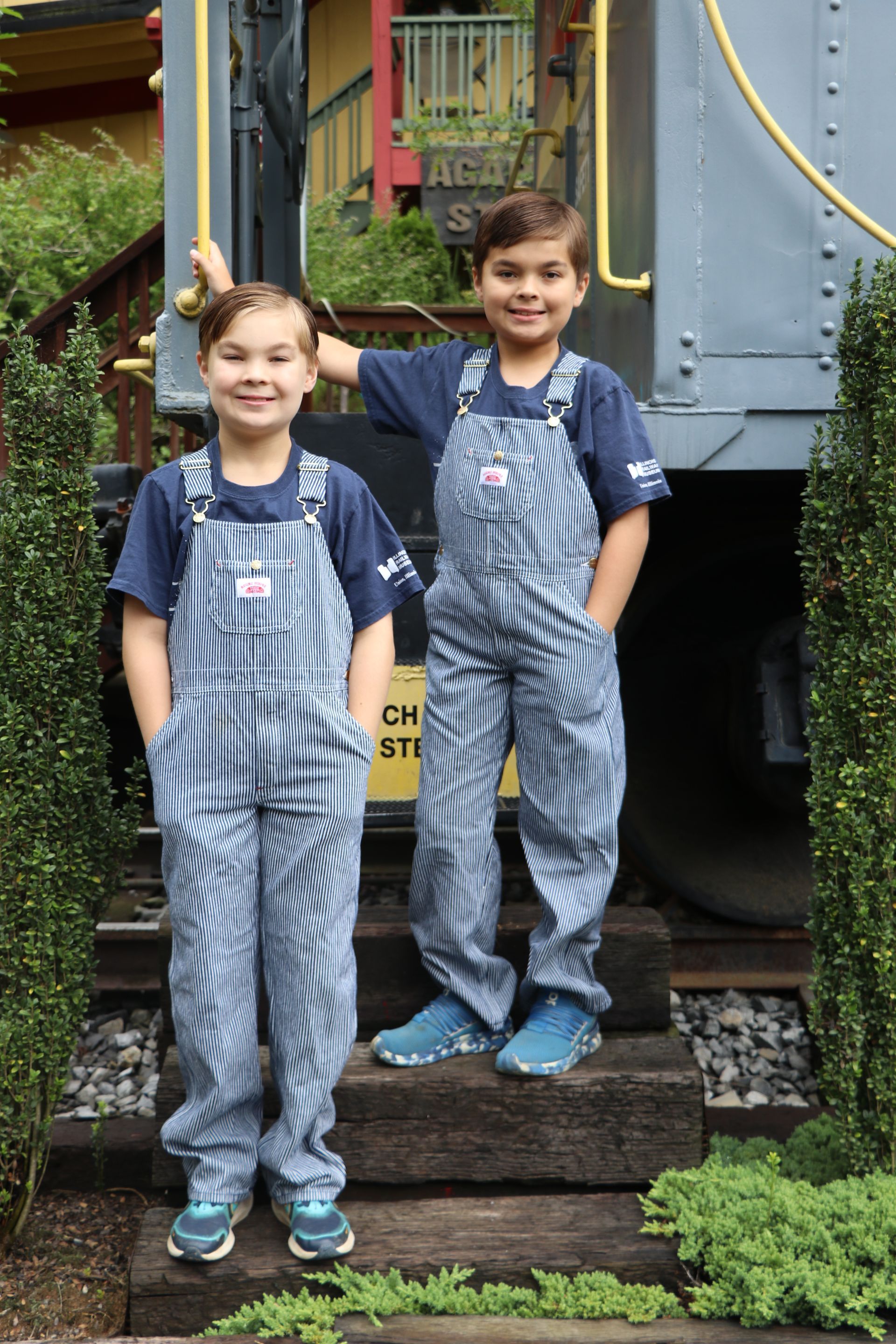 Two boys in striped overalls and blue shirts stand in front of a train.