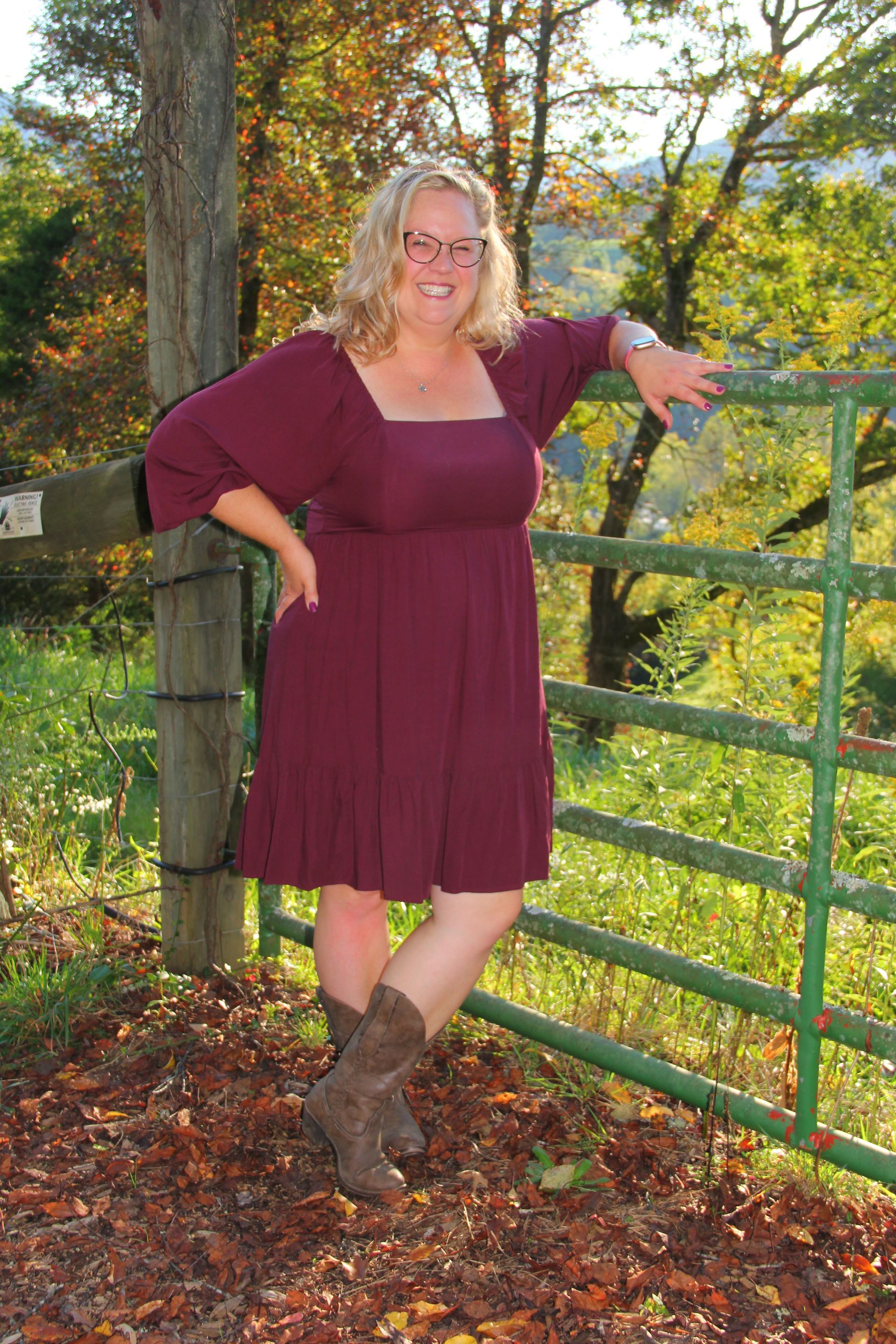 Woman in burgundy dress and cowboy boots leans on a green gate outdoors, smiling.