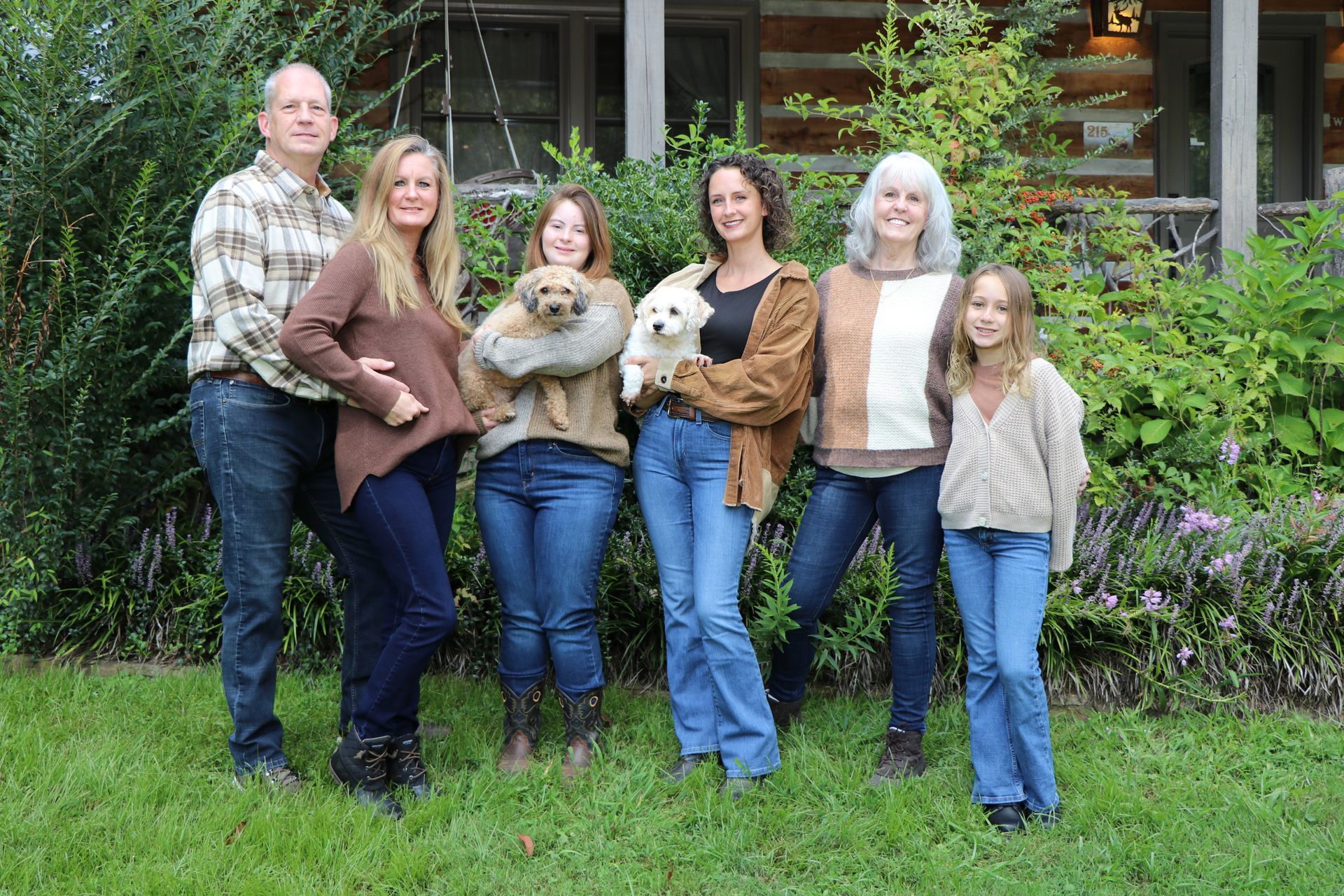 Family poses in front of a cabin, holding two small dogs. Green grass and foliage surround them.