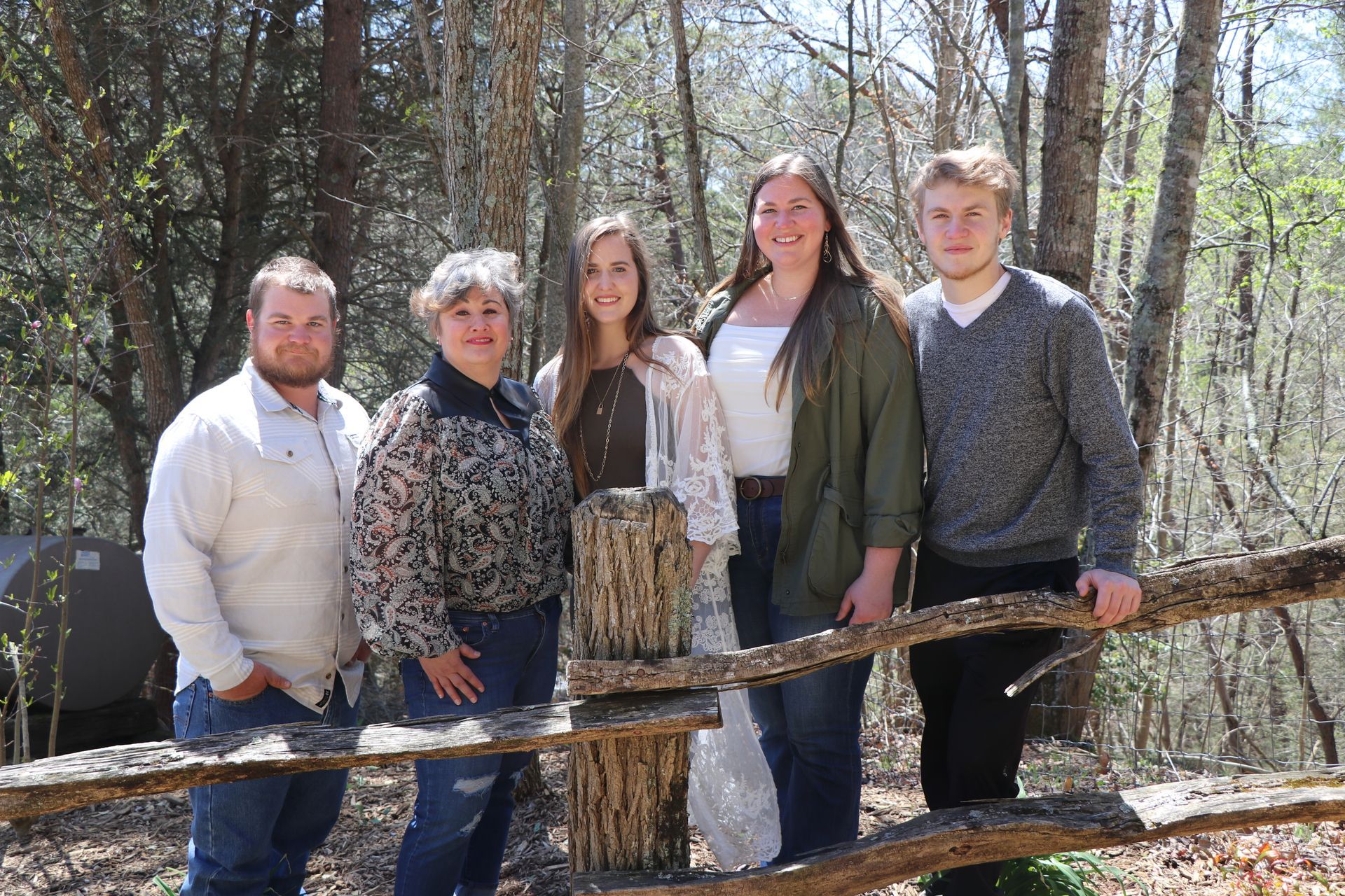 Group of five people standing behind a wooden fence, smiling outdoors amongst trees.