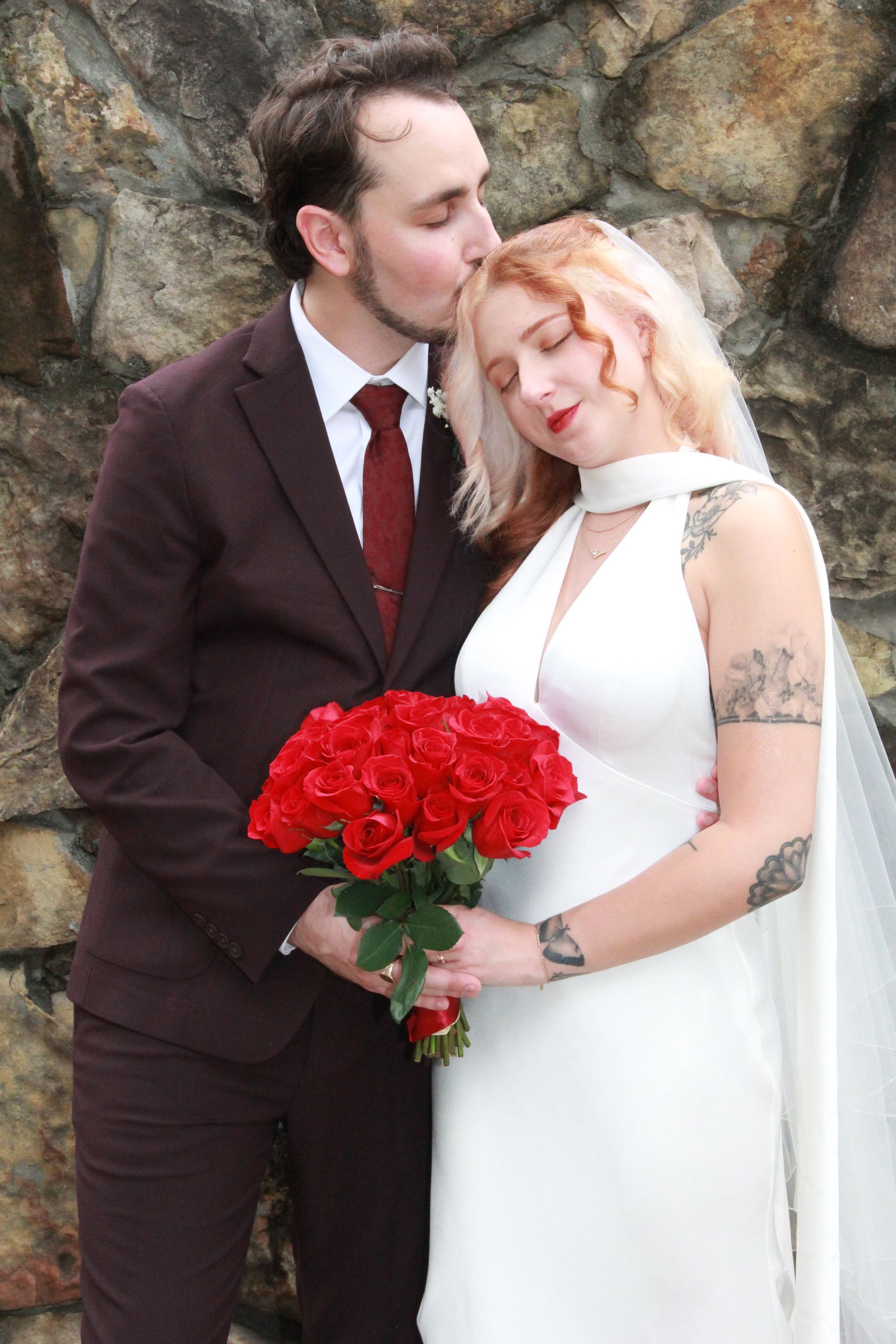 Groom kisses bride's forehead; holding bouquet of red roses, against a stone wall.
