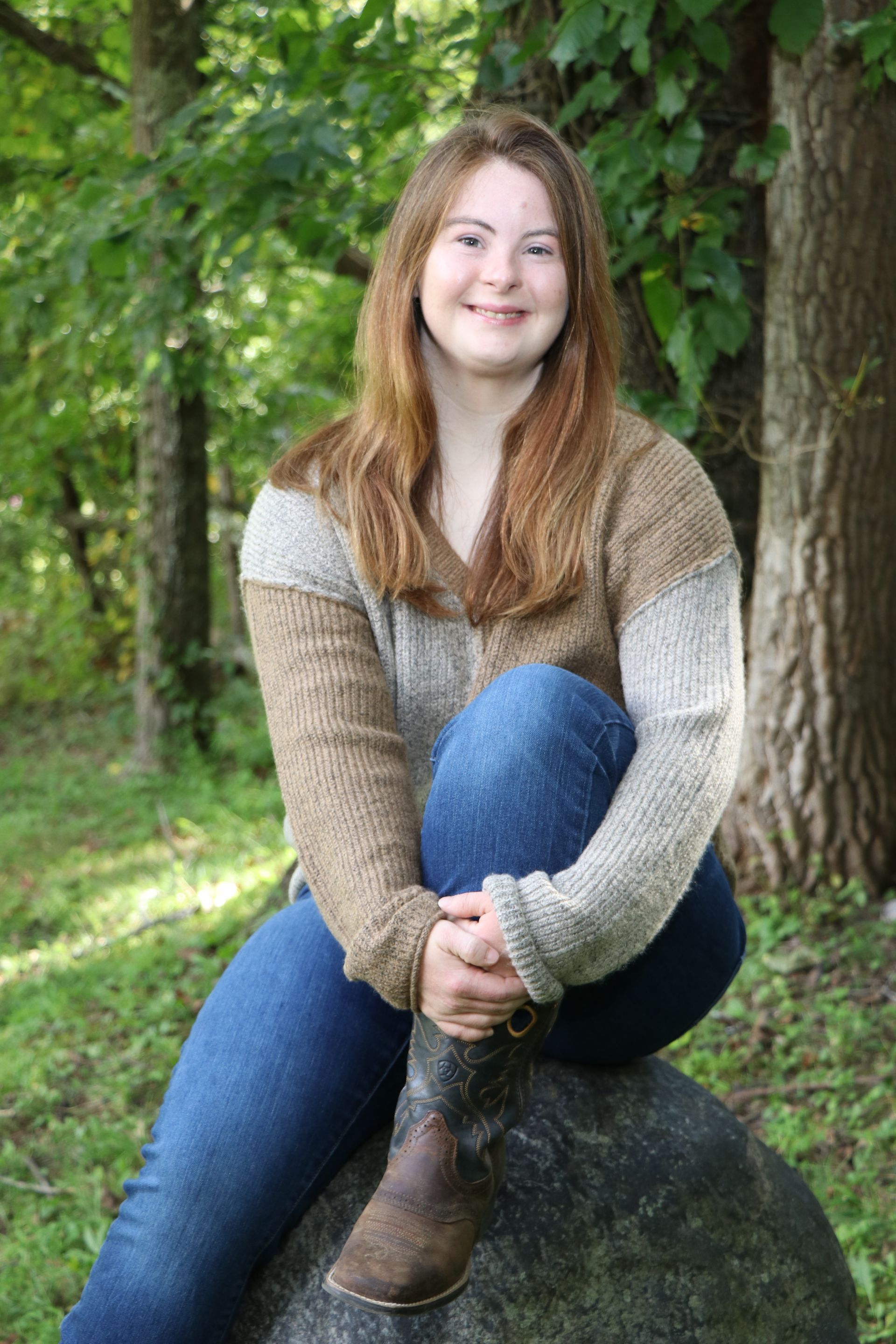 Woman with Down syndrome in sweater and jeans, smiling, sitting on a rock in a wooded area.