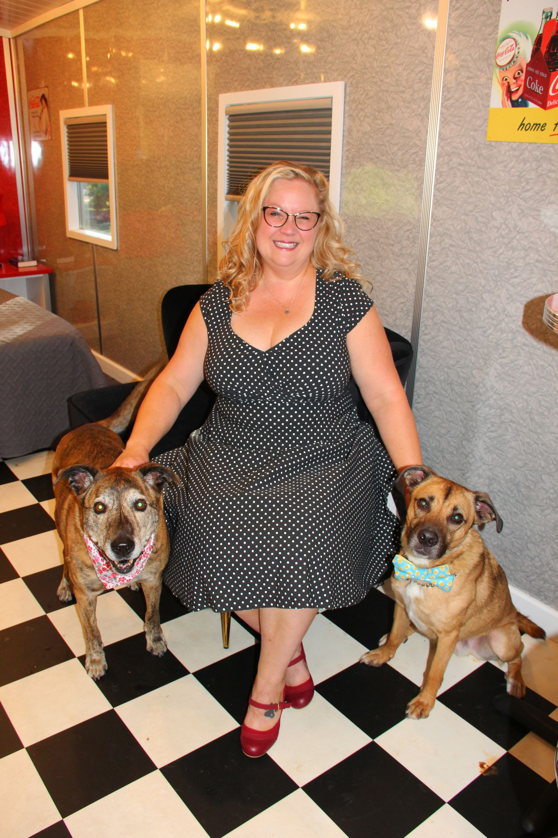 Woman in polka dot dress sits with two dogs on checkered floor.