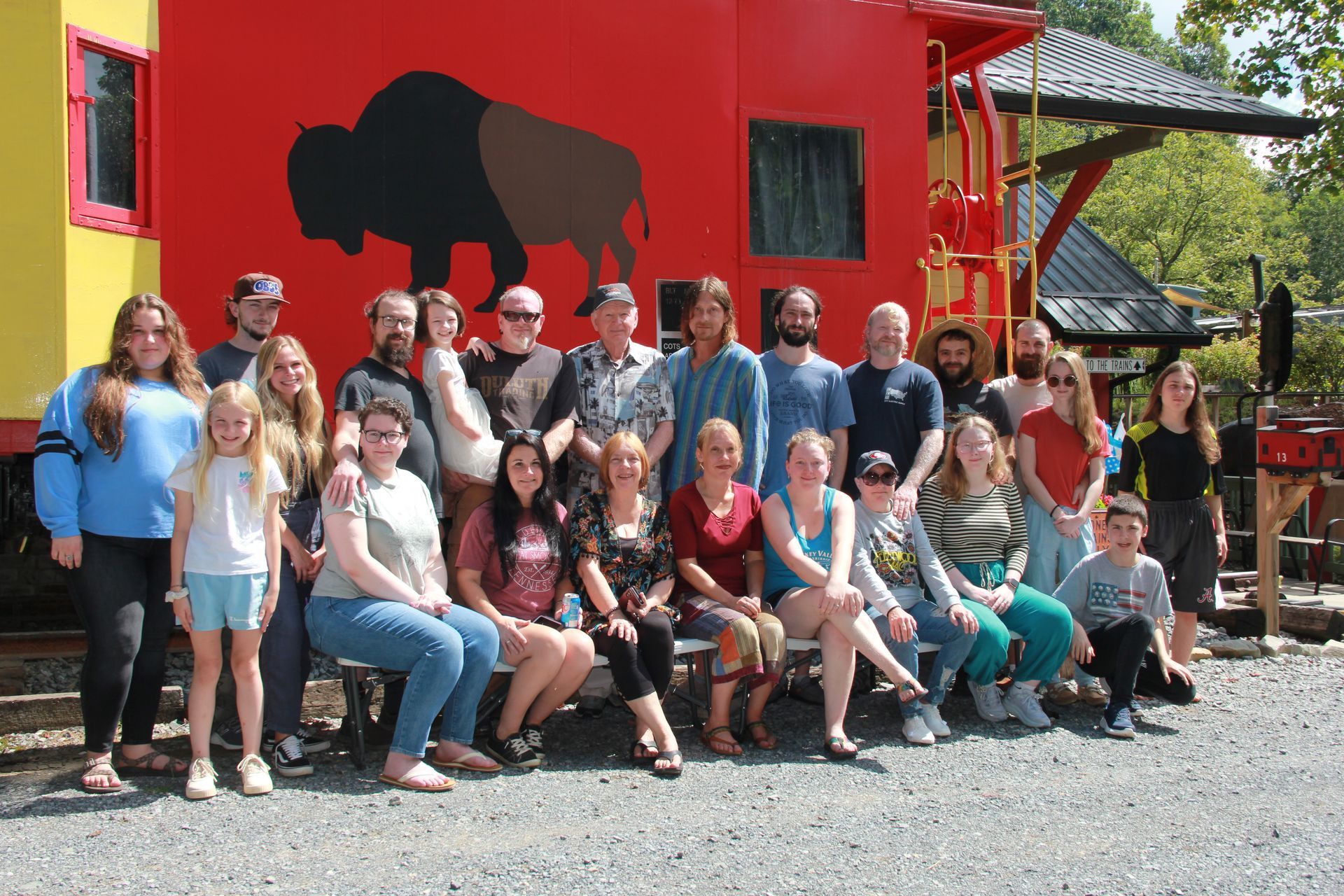Group of people posing in front of a red caboose with a bison silhouette; outdoor setting.
