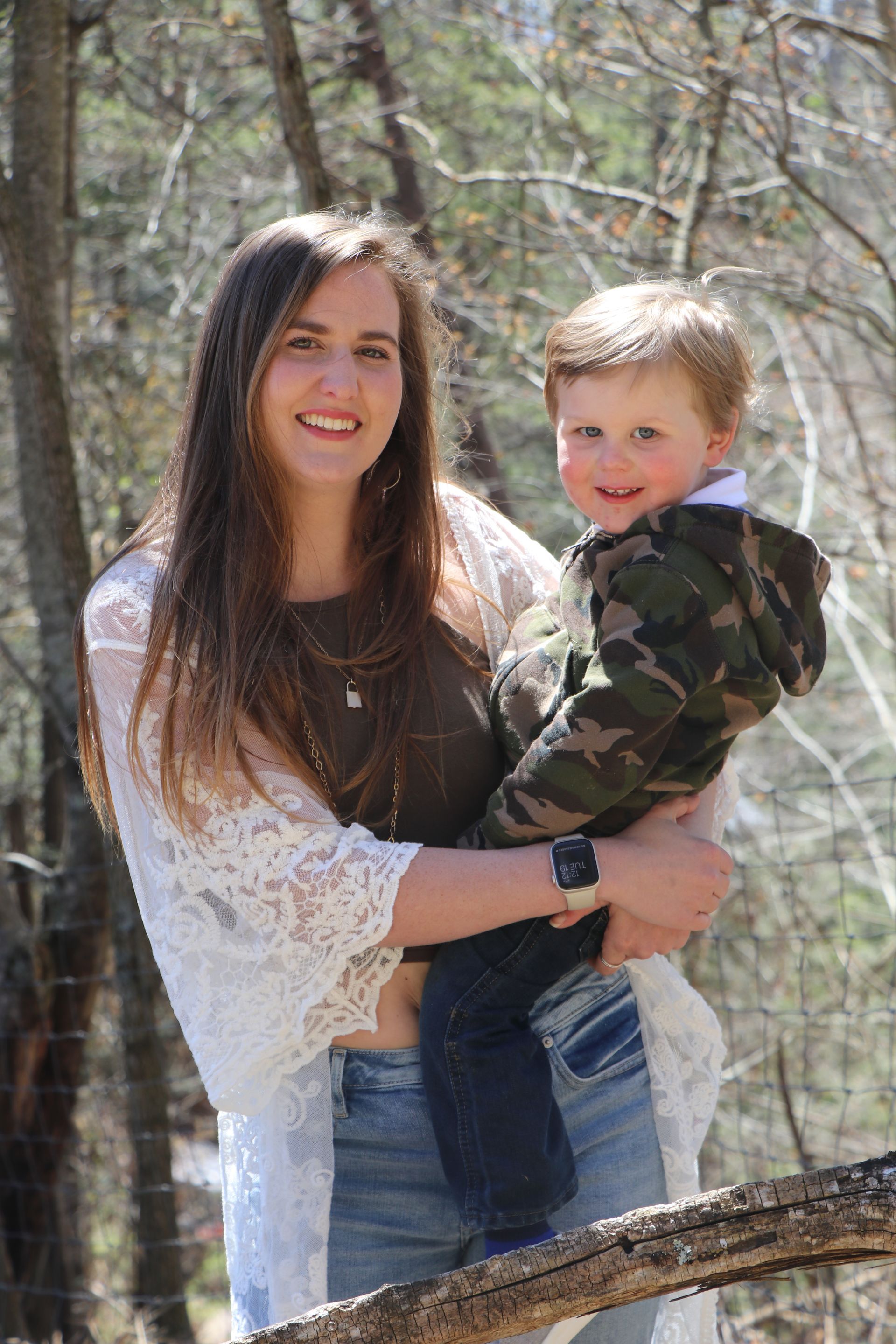 Woman with long brown hair holds a smiling young child in a forest. Both are outdoors, smiling.