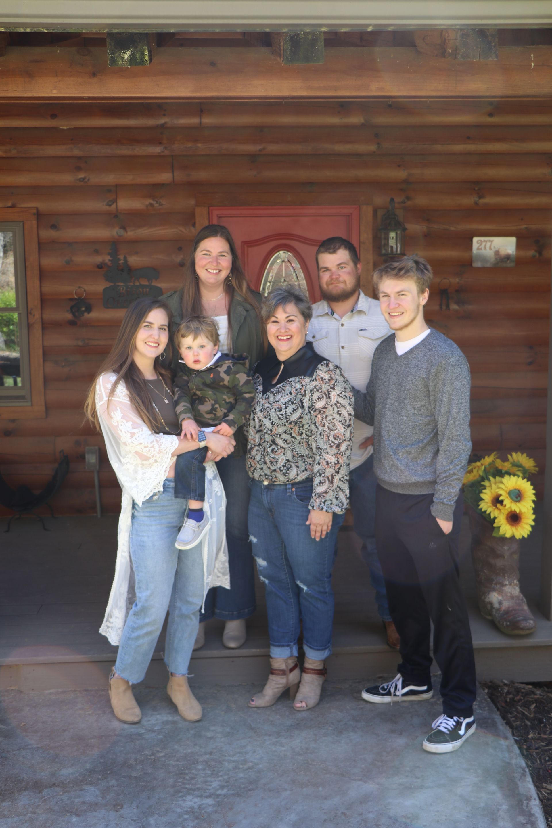Family of seven poses in front of a log cabin with a red door.