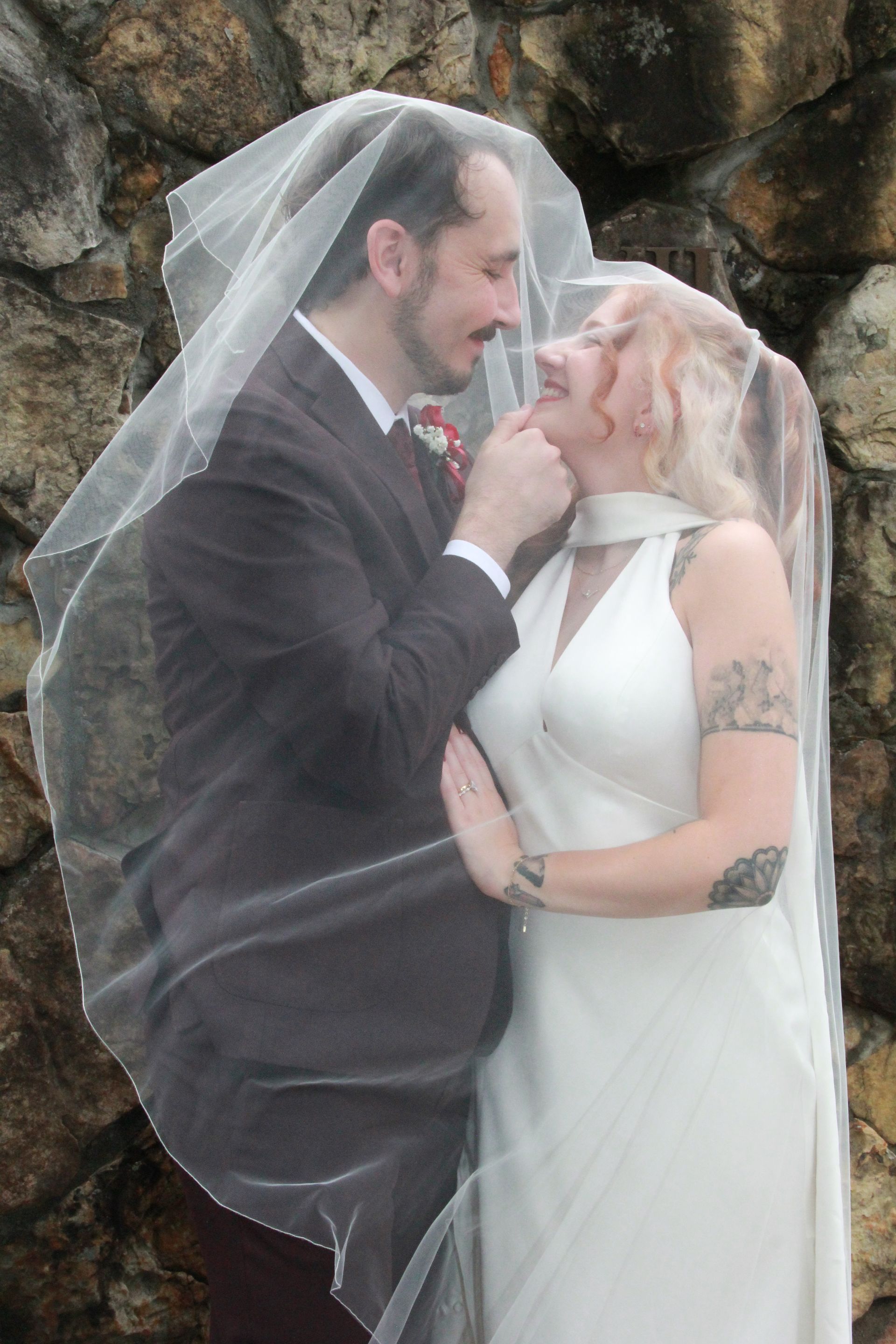Bride and groom smiling under a veil, against a stone wall. Groom touches bride's chin.