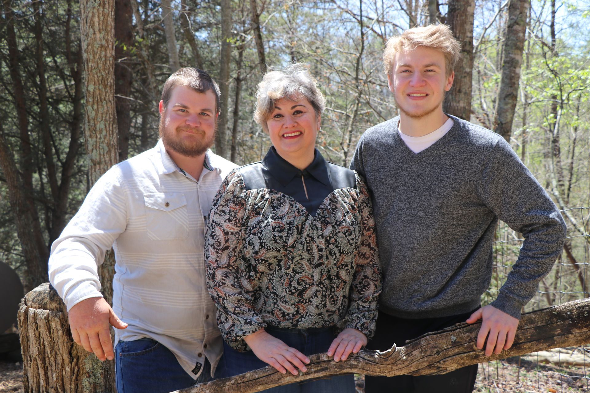 Family posing outdoors, smiling. Mother in patterned shirt between two men, leaning on a log in a forest.