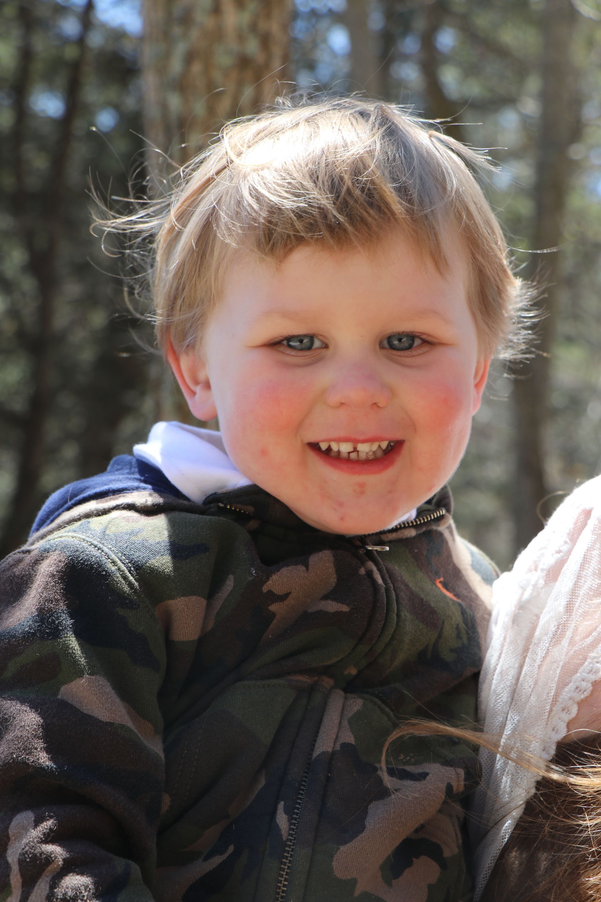 Young child smiling with rosy cheeks, wearing a camouflage jacket outdoors.