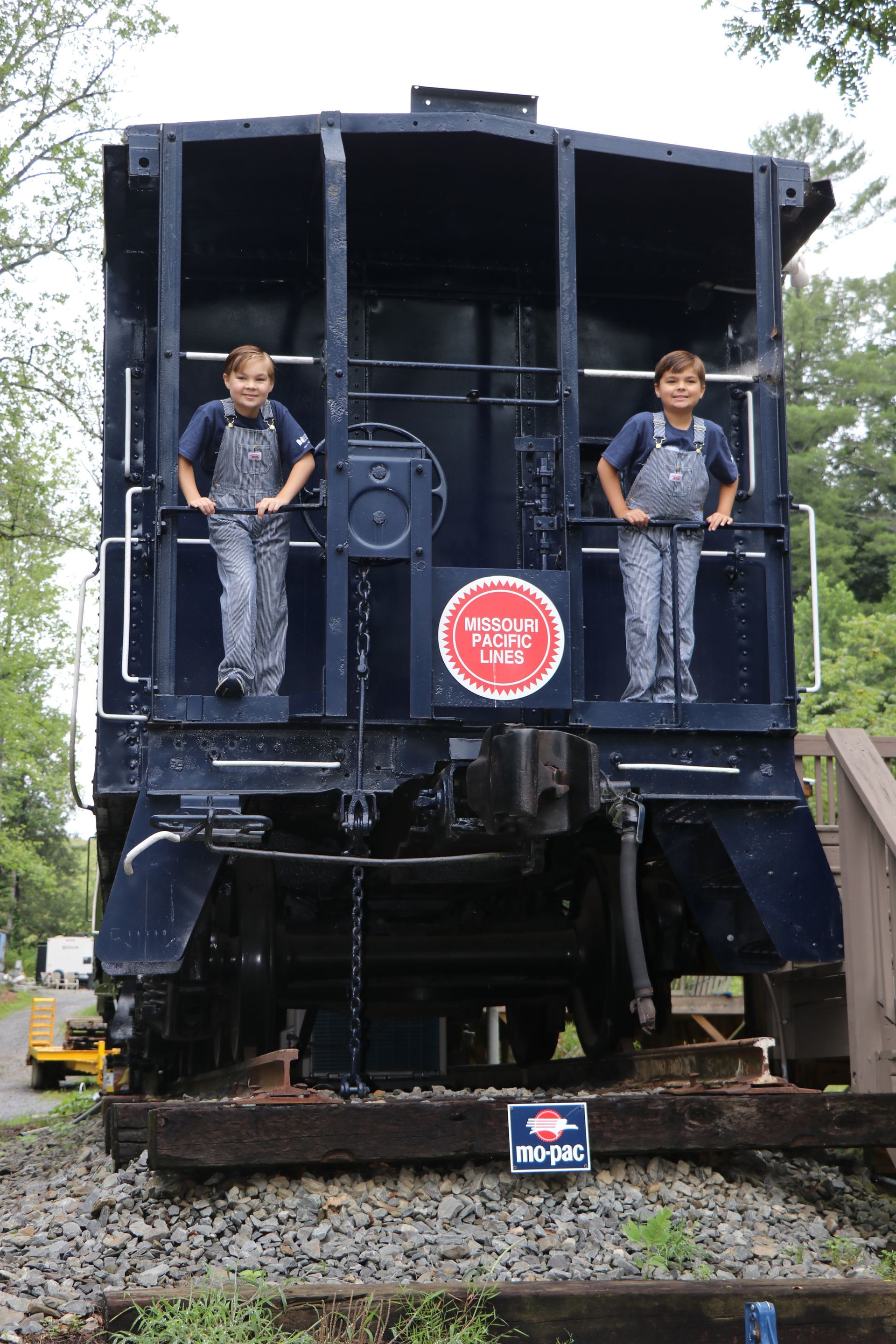 Two children in overalls standing in a dark blue train caboose, smiling, with a railroad sign in front.