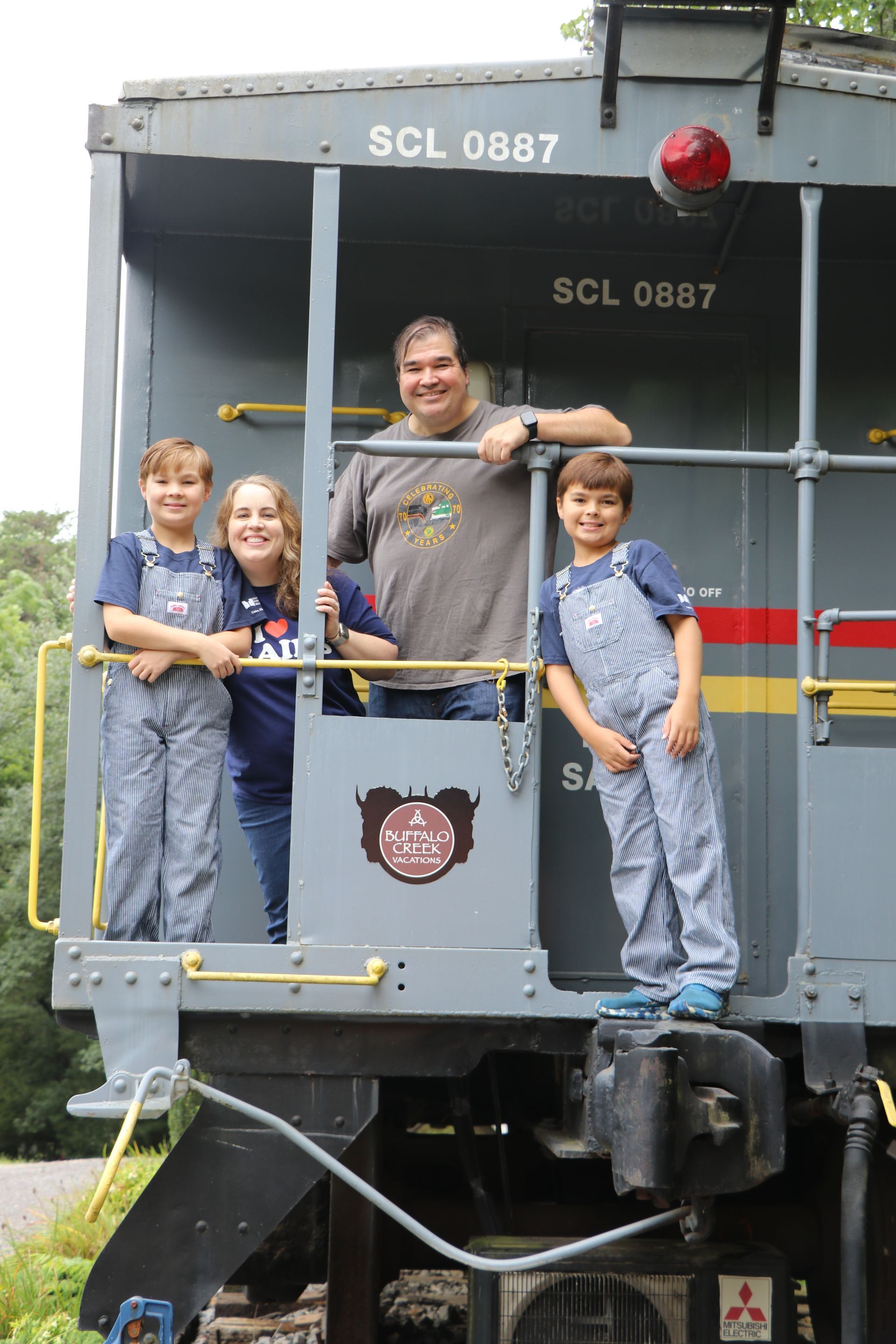 Family poses in a gray train car. The children wear blue overalls, and the man wears a gray shirt.