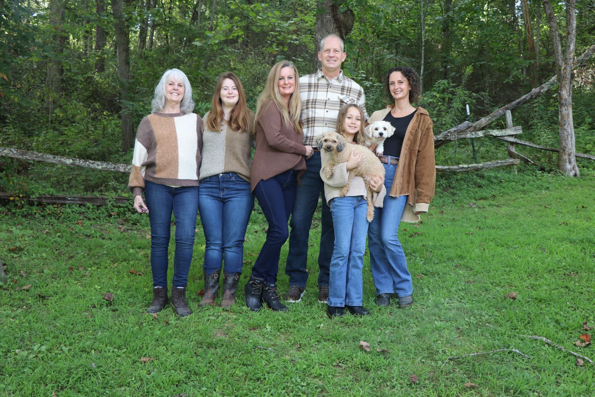 Family of six stands outside on grass near a wooded area; holding a small dog.