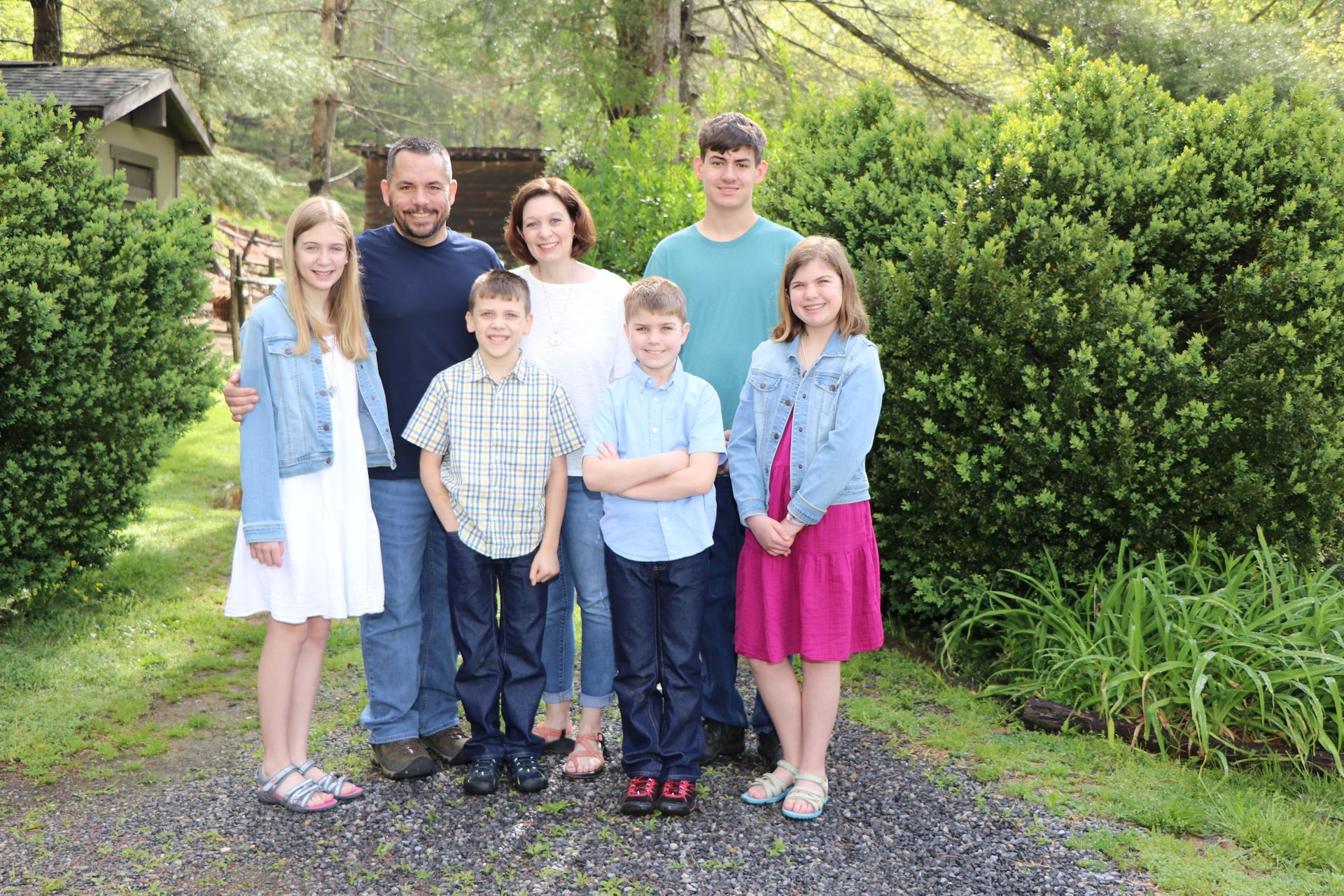 Family of seven poses outdoors. Adults, children, smiles. Green foliage, gravel path.