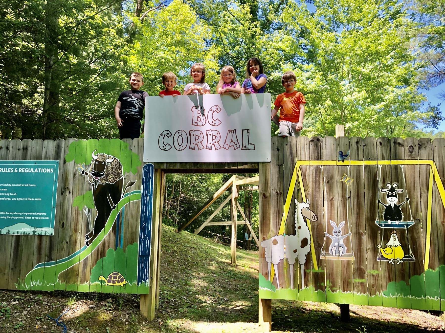 Children on fence above a wooden structure with "The Corral" sign. Cartoon animal cutouts painted on fence.