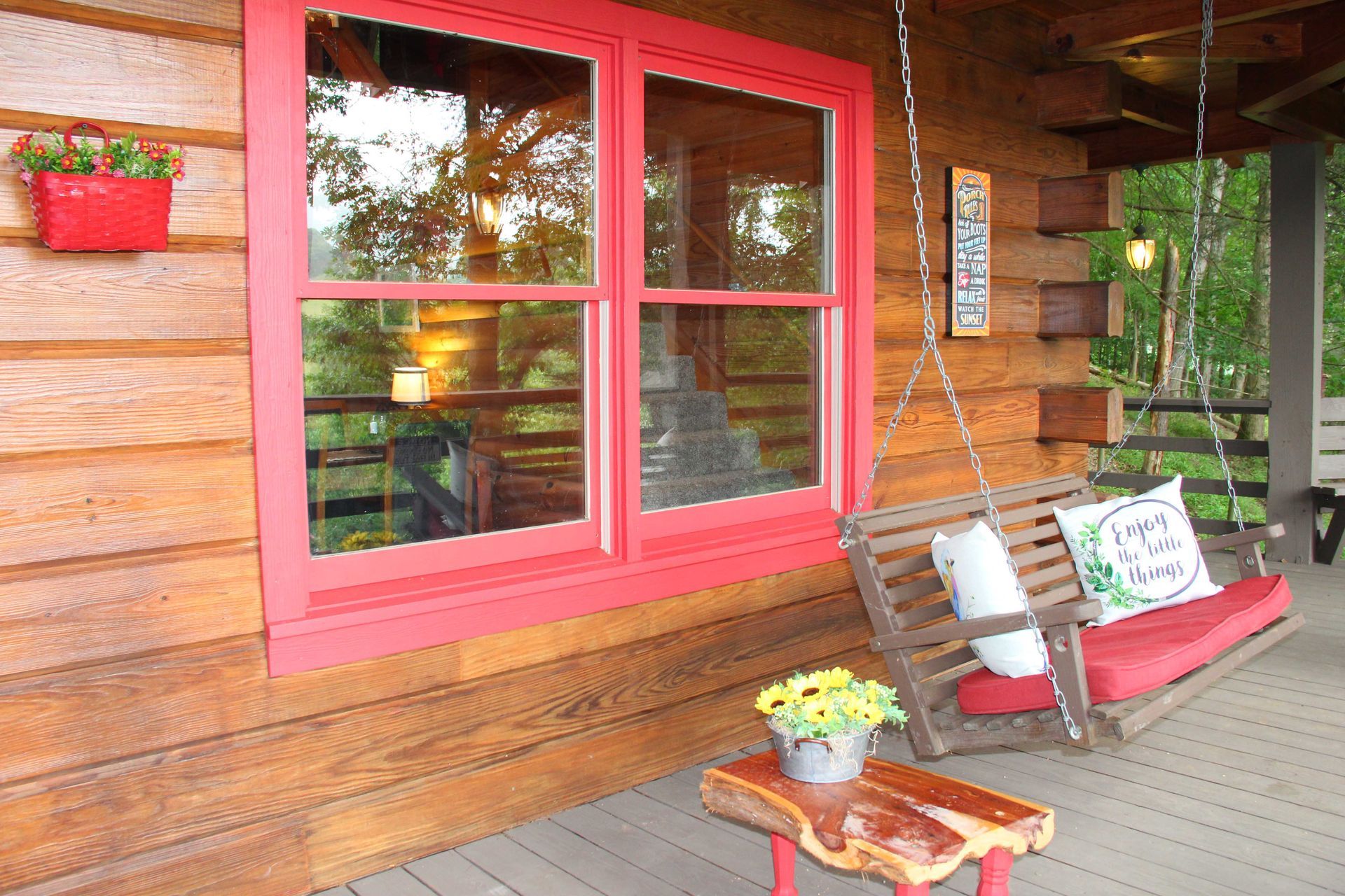 Log cabin porch with a red-framed window, swing, and a small table with sunflowers.