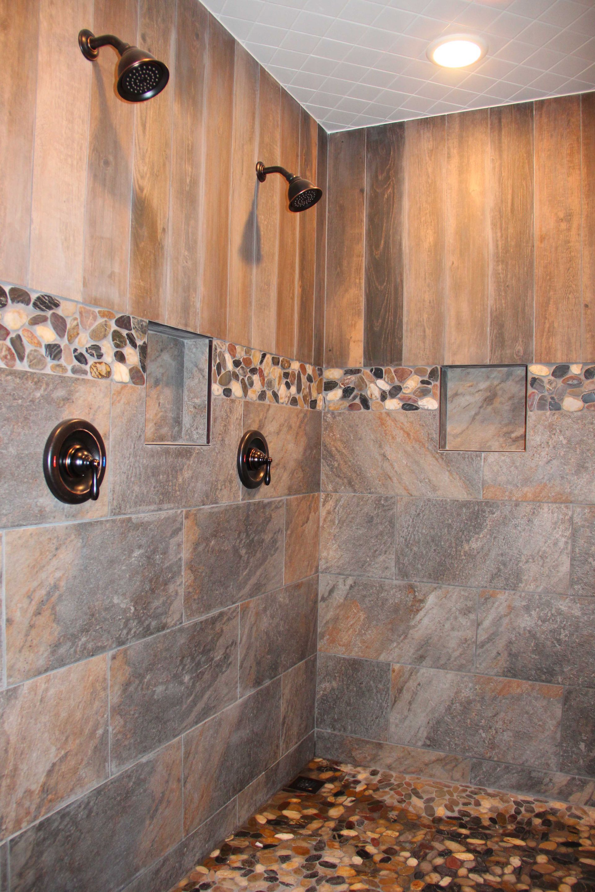 Shower with brown and gray stone tiles, pebble floor, two shower heads, and built-in soap niches.