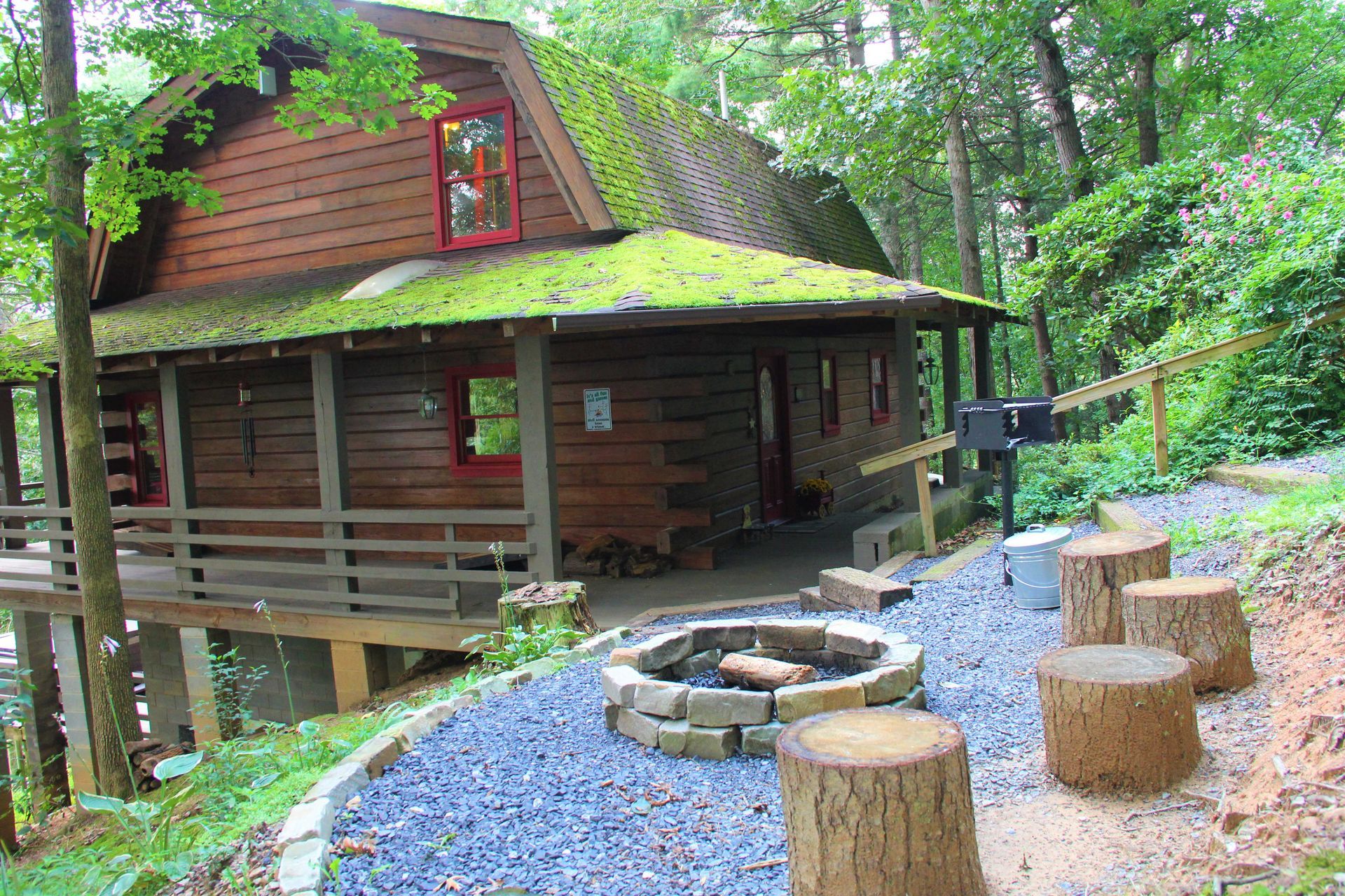 Log cabin with mossy roof, porch, fire pit, and tree stump seating in a wooded setting.
