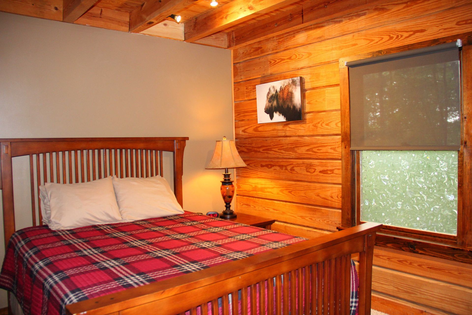 Bedroom with wooden walls, a plaid comforter, and a window with a partially drawn shade.