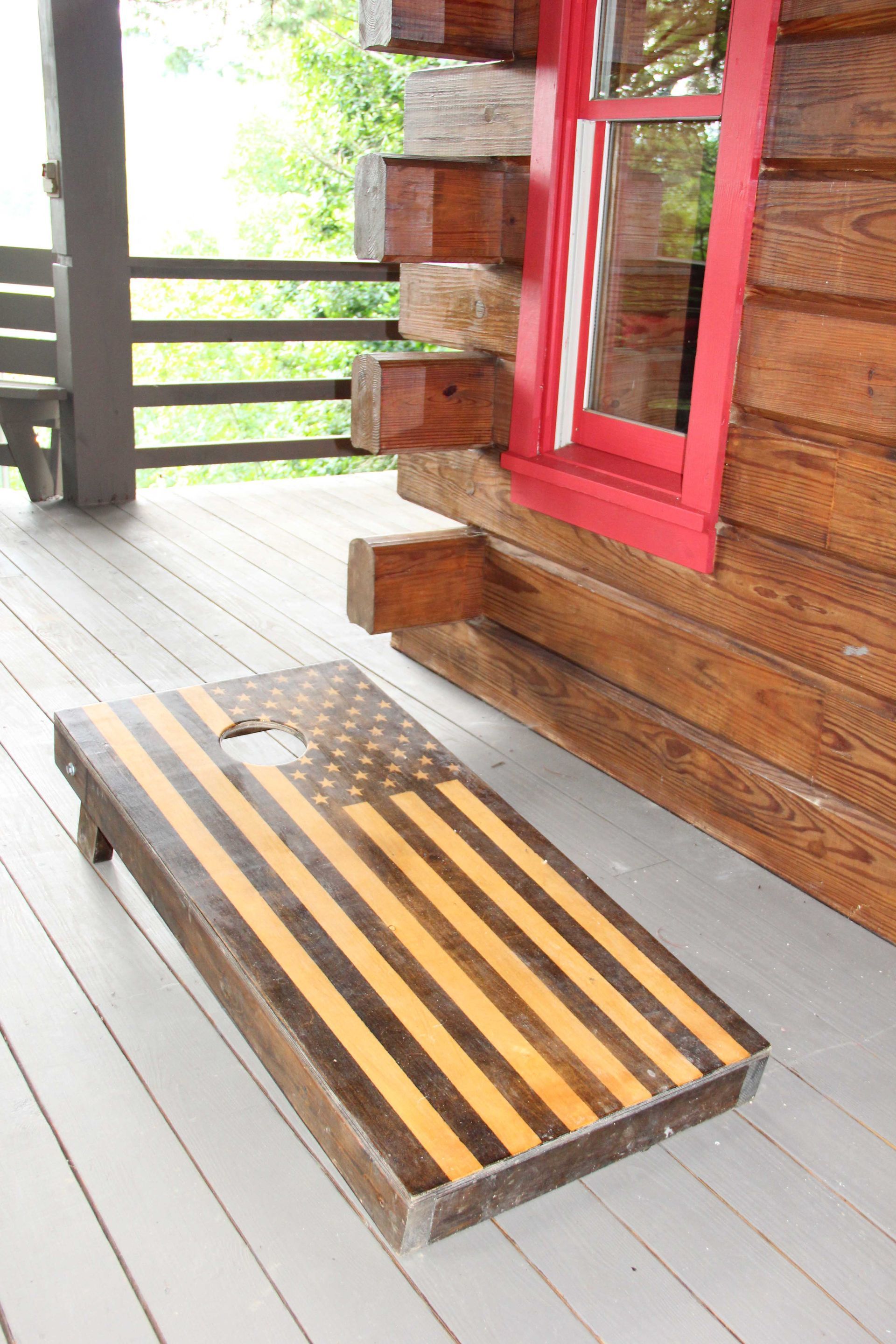 Cornhole board with American flag design on porch of log cabin.