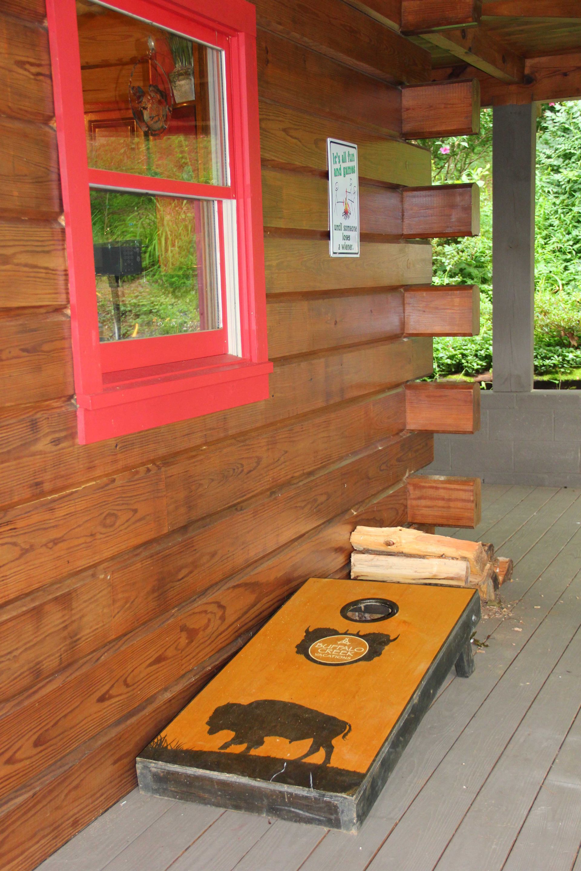 Log cabin exterior with a red-framed window and a cornhole board with a buffalo graphic on a porch.