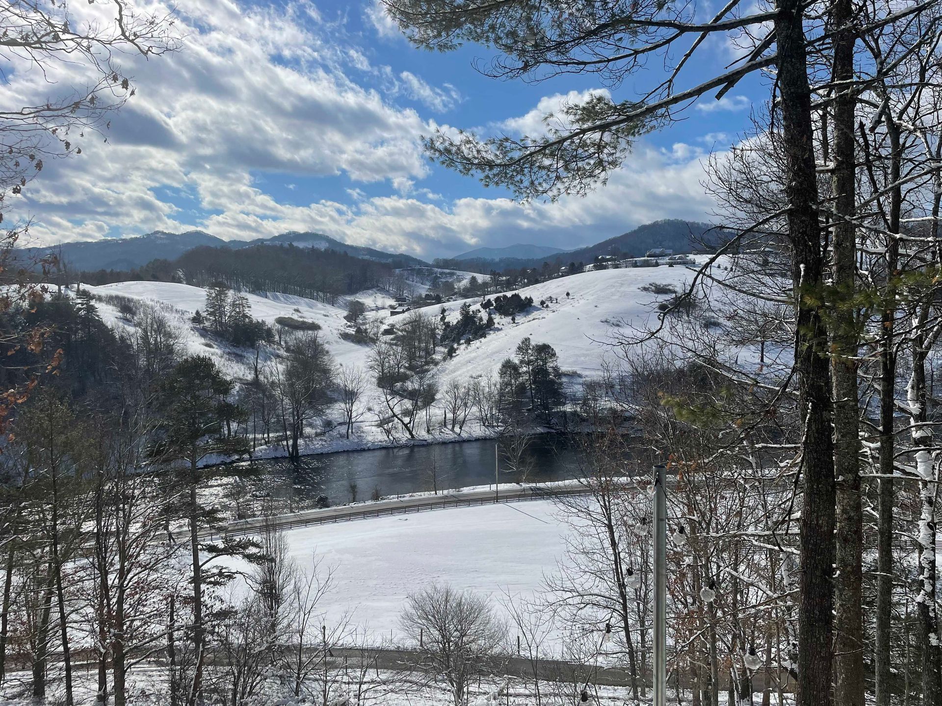 Snow-covered mountain landscape with river and cloudy blue sky, trees in foreground.