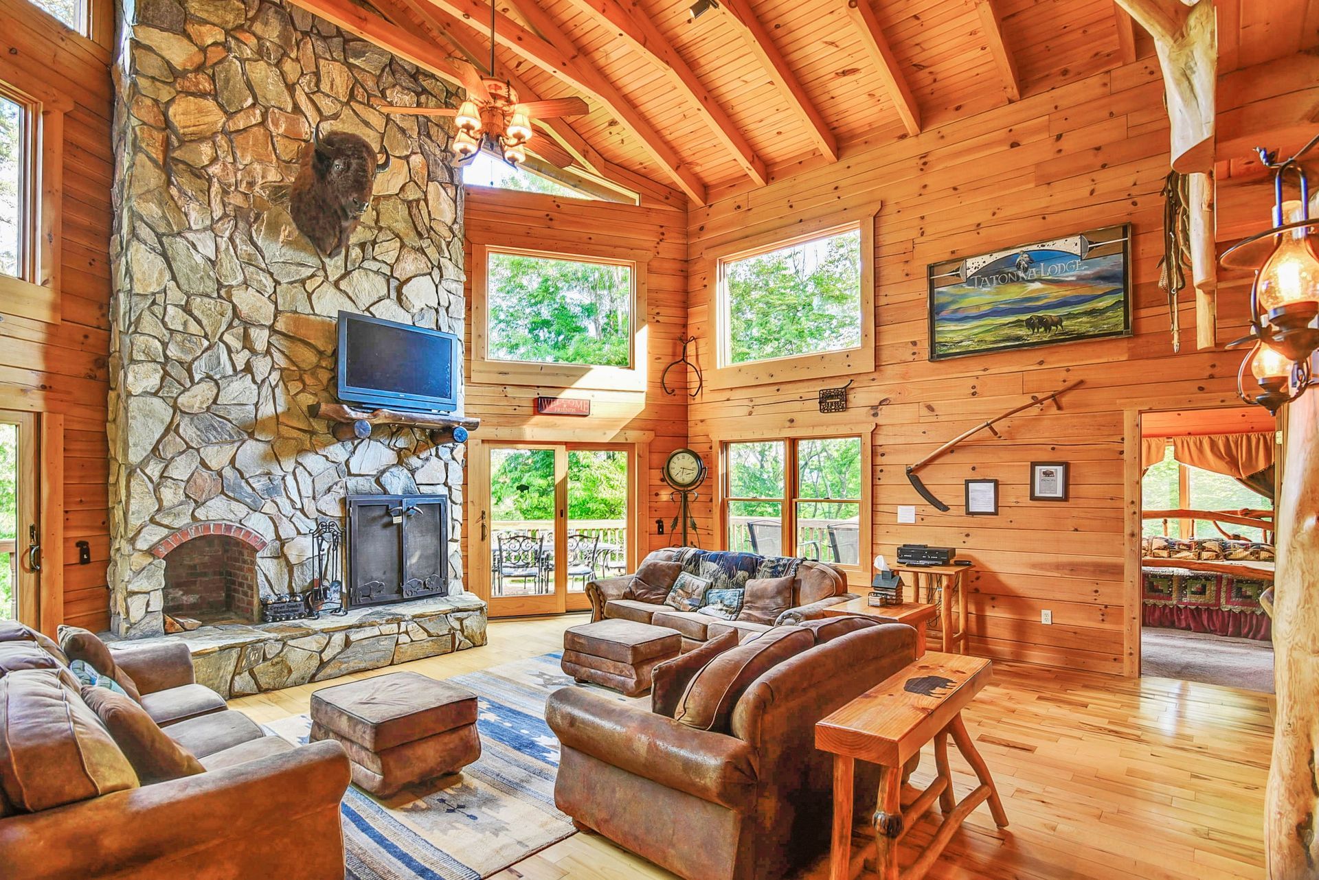 Spacious log cabin living room with stone fireplace, leather furniture, and large windows overlooking greenery.