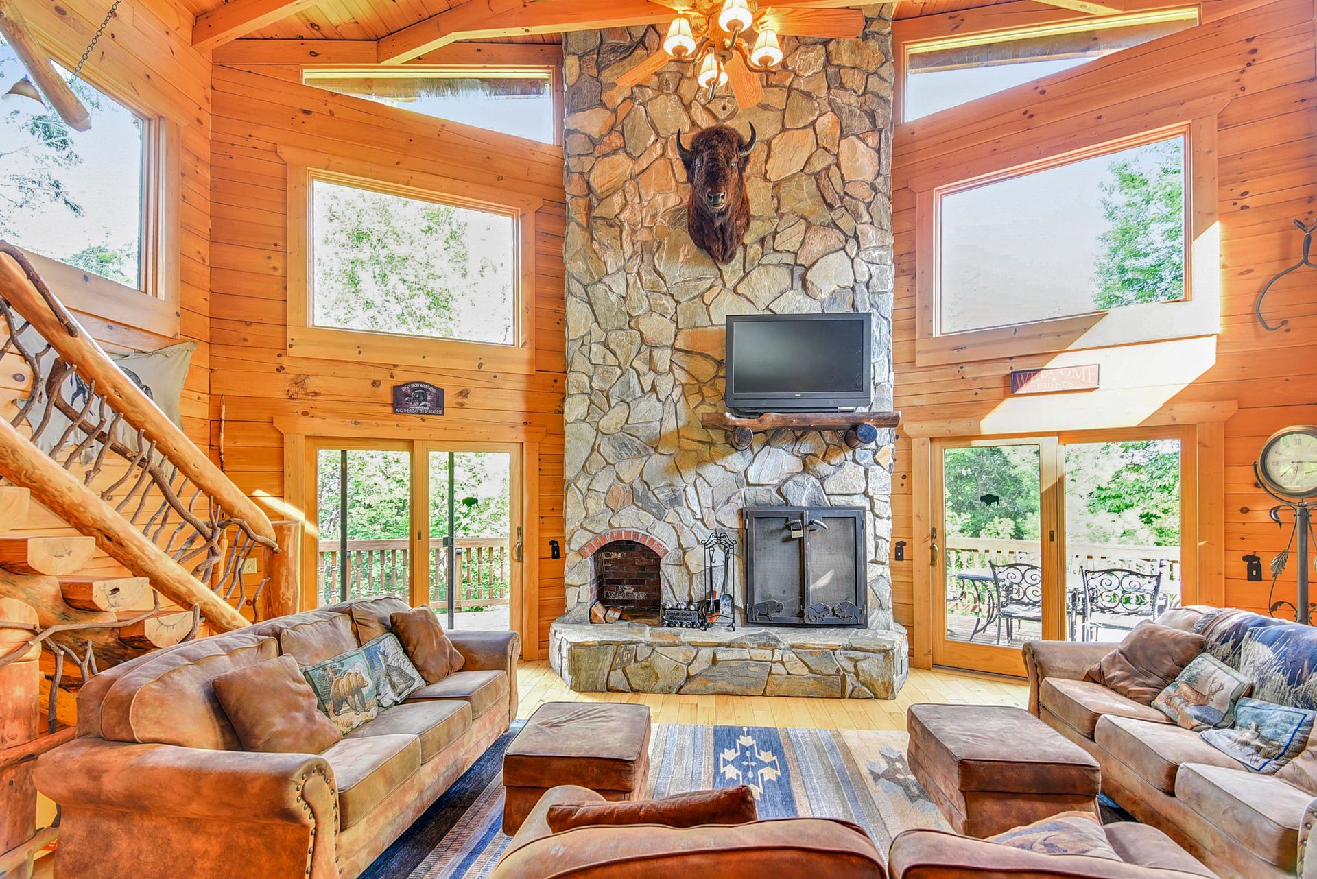 Cozy log cabin living room with stone fireplace, two sofas, windows, and wooden staircase.