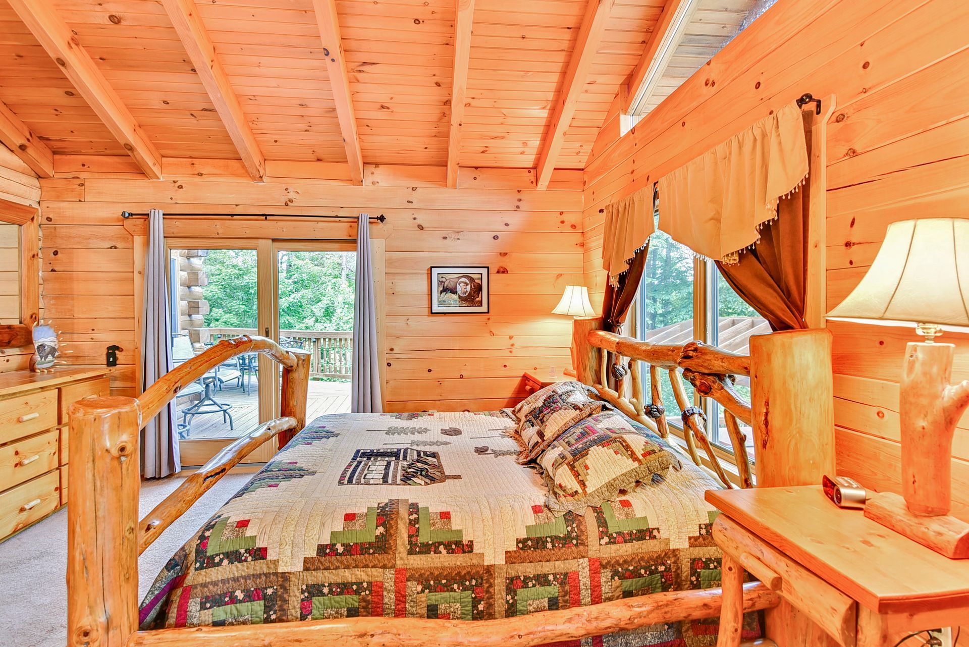 Bedroom in a cabin, wooden walls and ceiling. Queen bed with quilt, bedside tables, and a balcony visible.