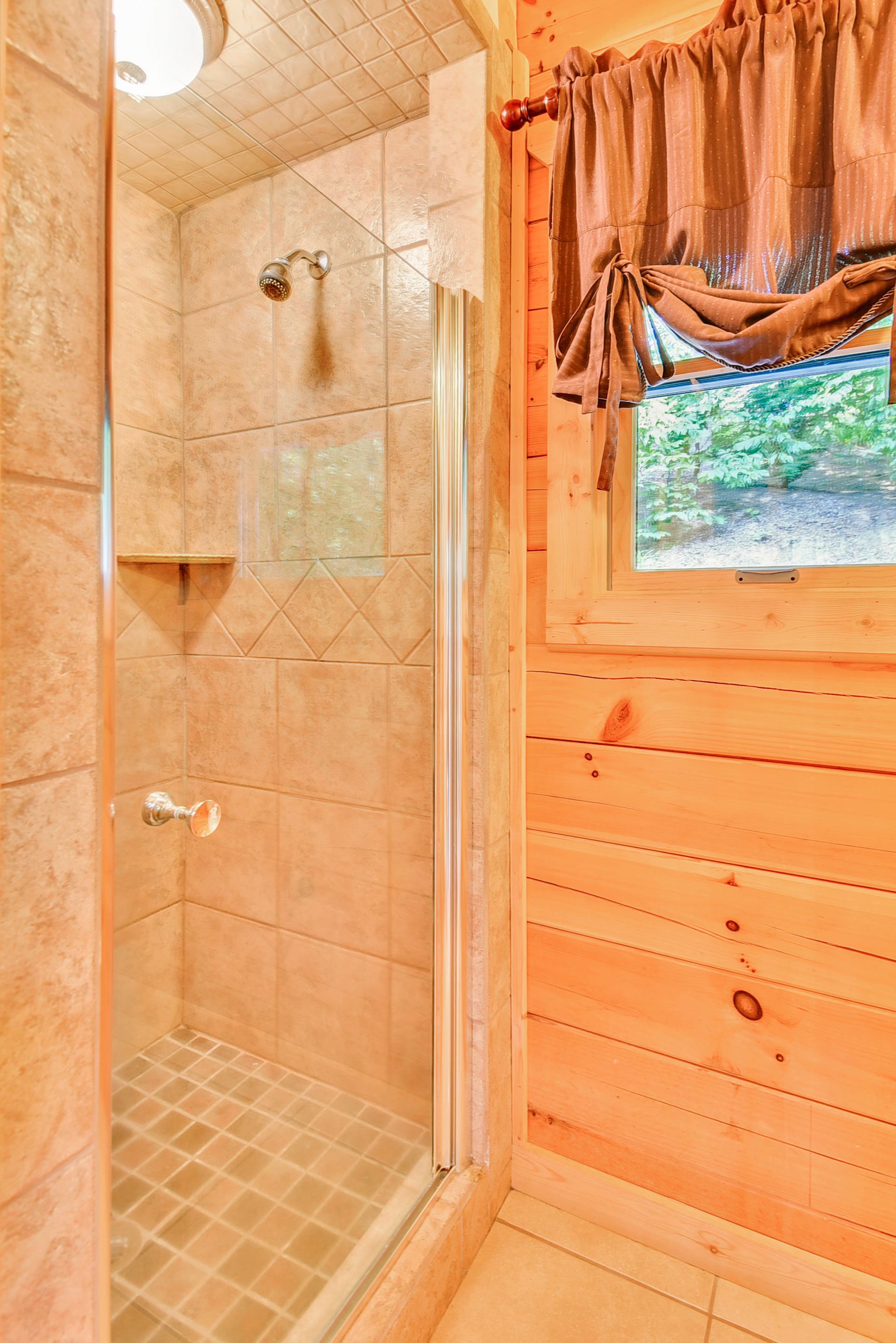 Small, beige-tiled shower in a wooden cabin bathroom. A window with a brown curtain is on the right.