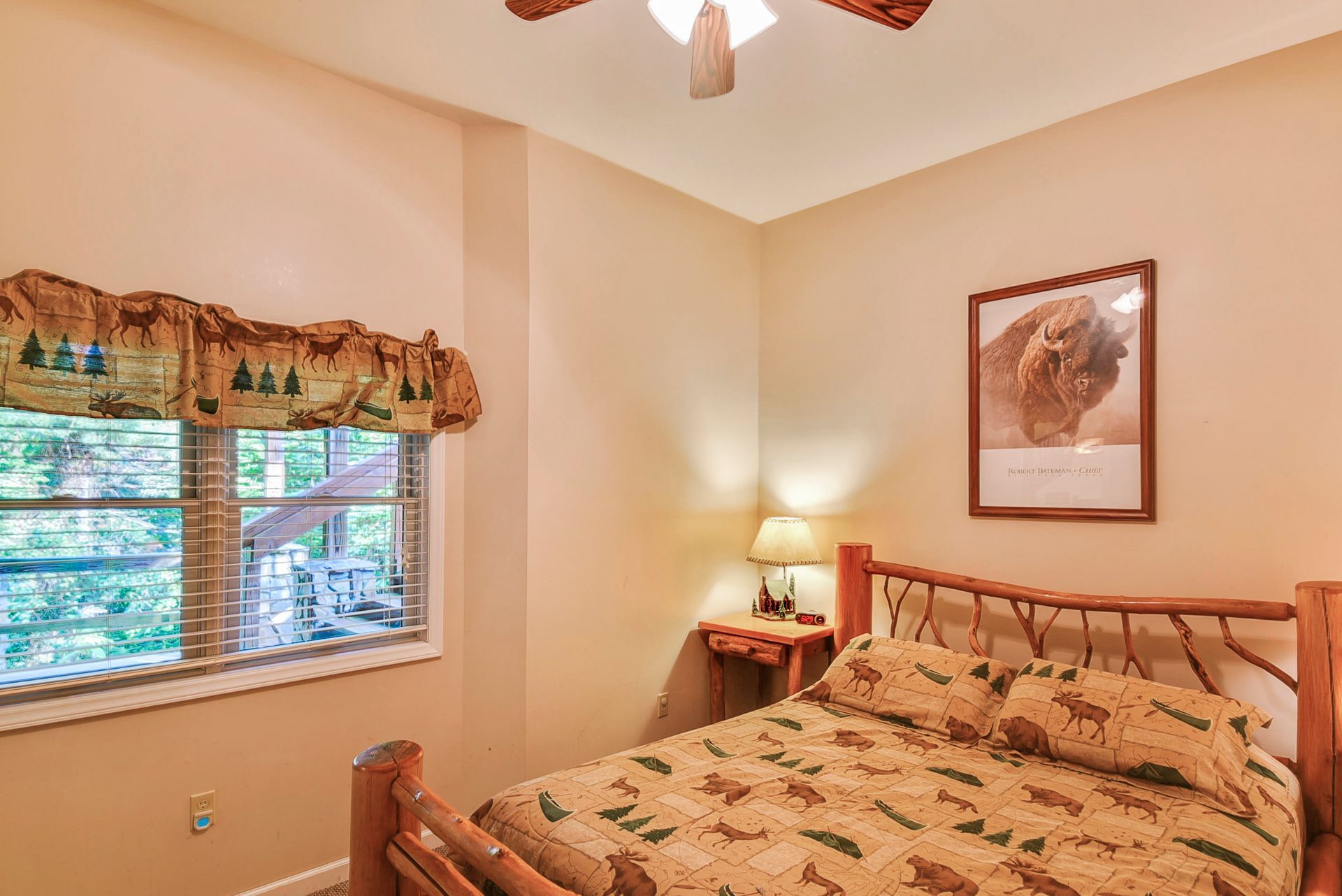 Bedroom with a rustic wooden bed, small table with a lamp, and a window with a valance.