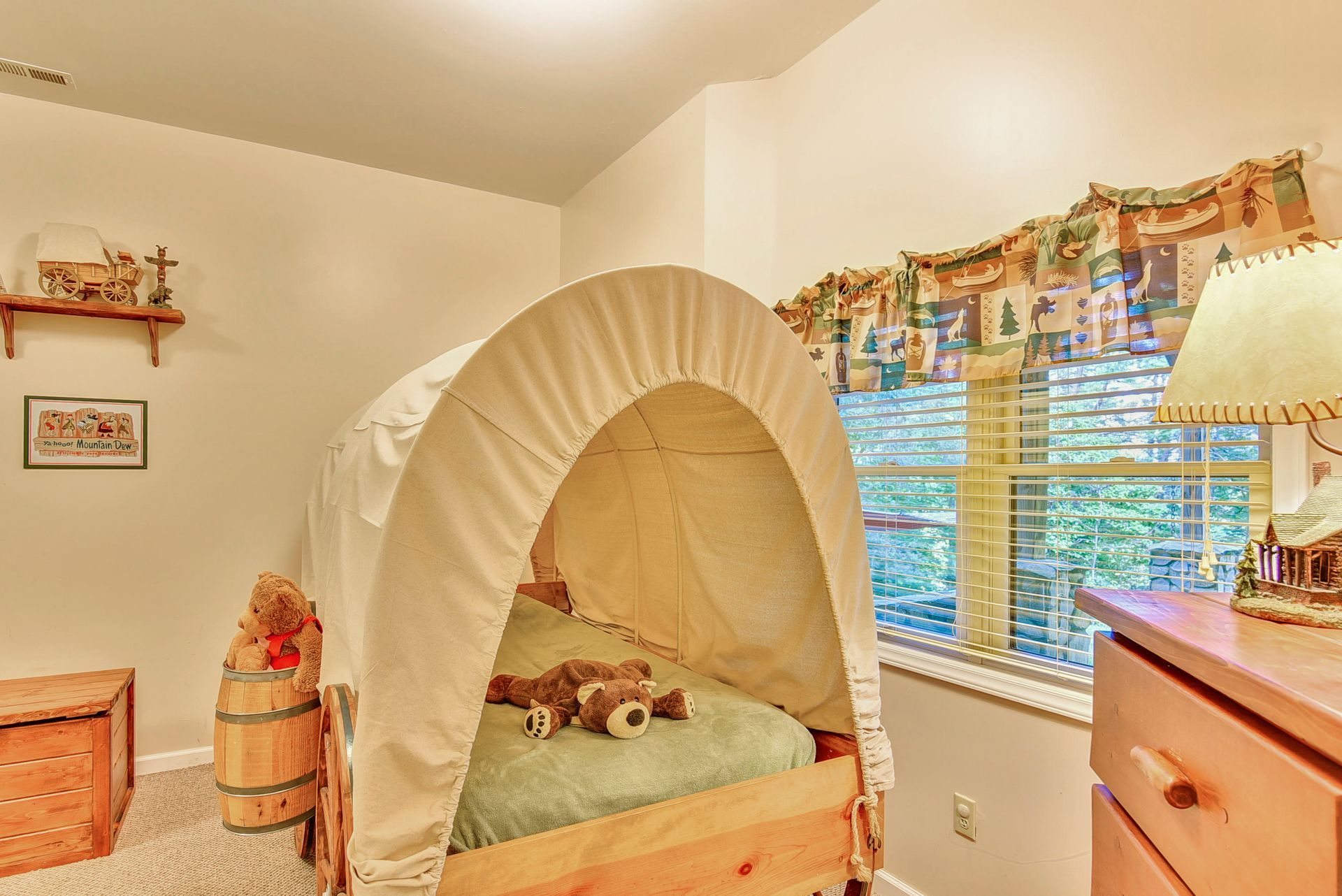 A child's bedroom with a covered wagon bed, chest, shelf, and window with a view of trees.