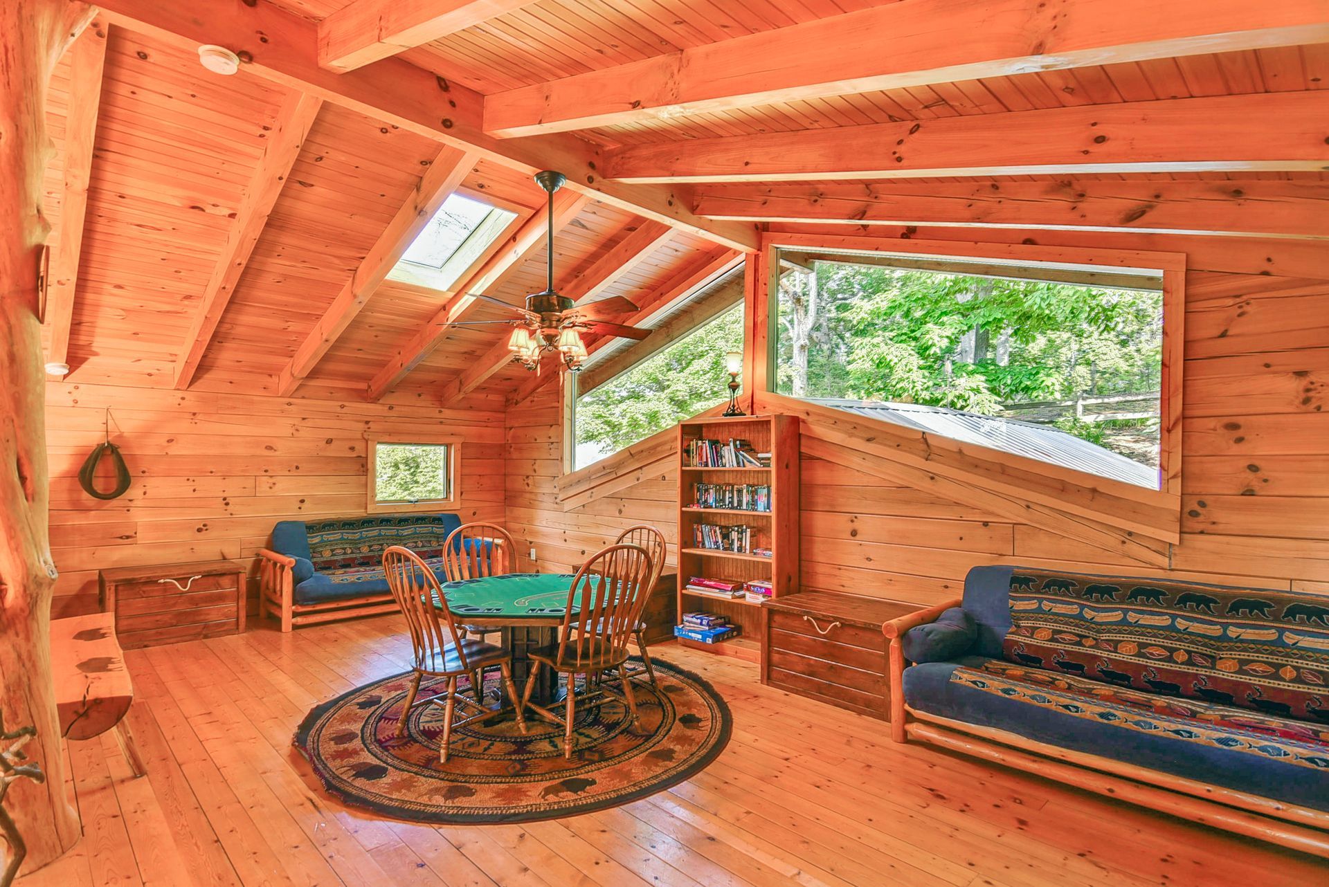 Wooden cabin game room with round table, sofas, and large windows overlooking trees.