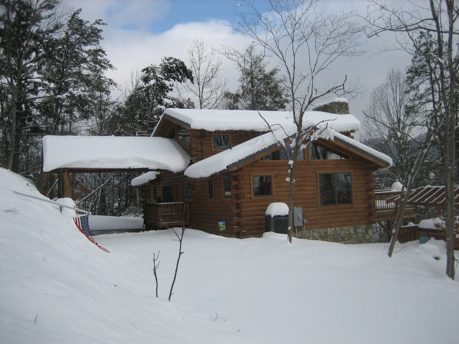 Log cabin in winter; snow-covered roof, yard, and trees. Brown logs, blue sky, and a covered porch are visible.
