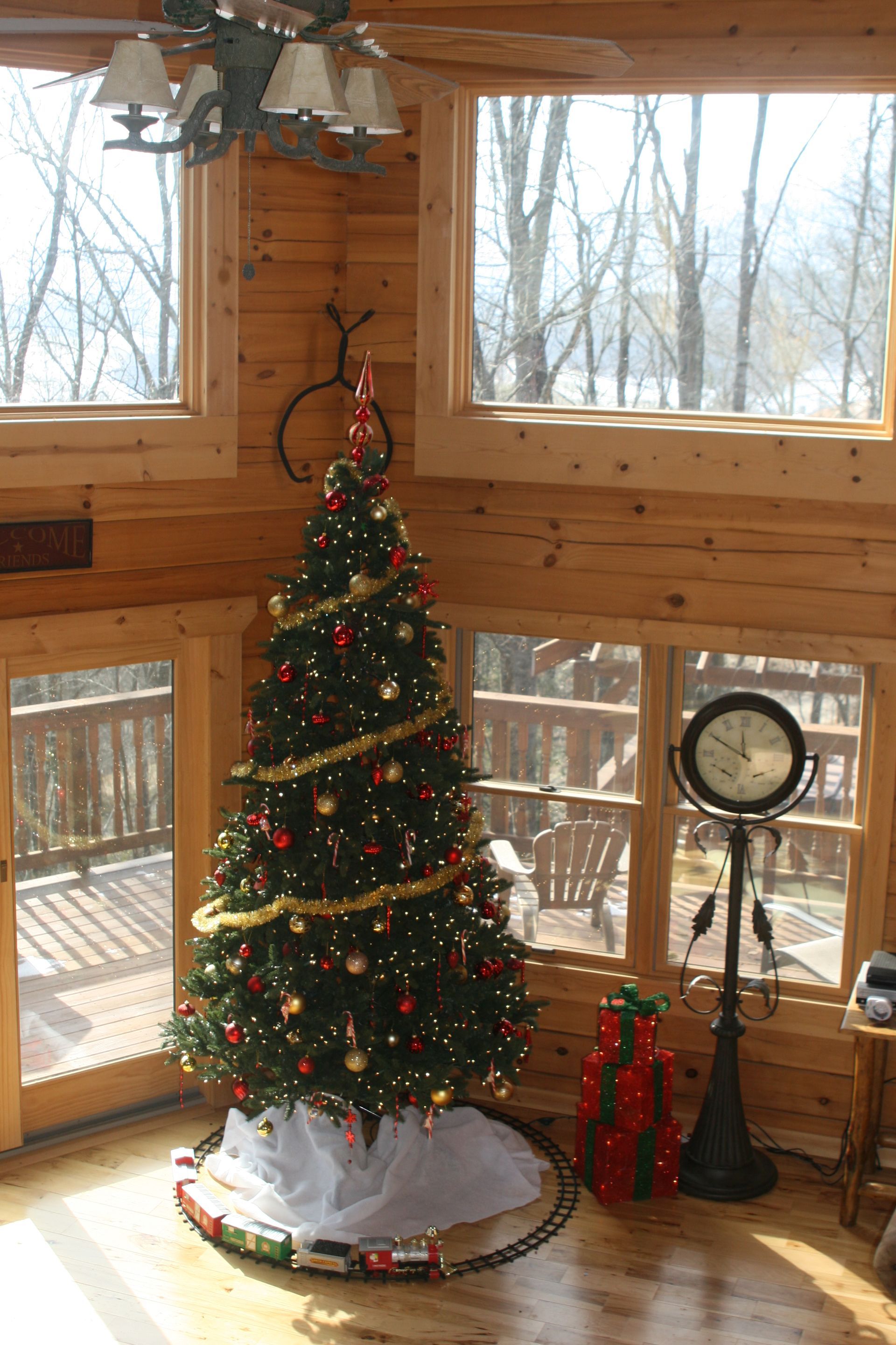 Christmas tree in a wooden cabin corner by large windows, garland, lights, train track, and decorative gifts.