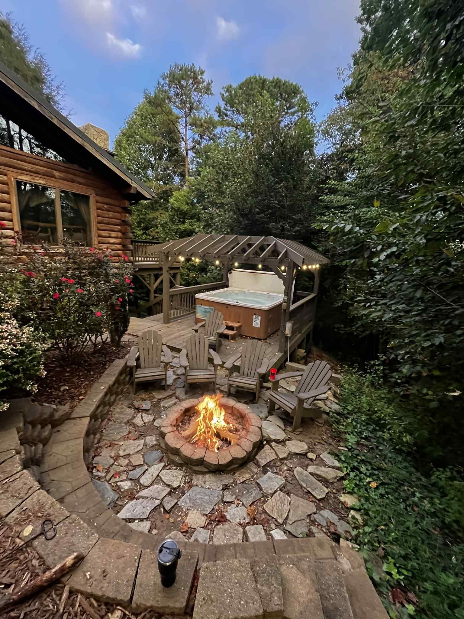 Cozy outdoor scene with a fire pit, hot tub, and log cabin. Evening setting, string lights, chairs, trees.