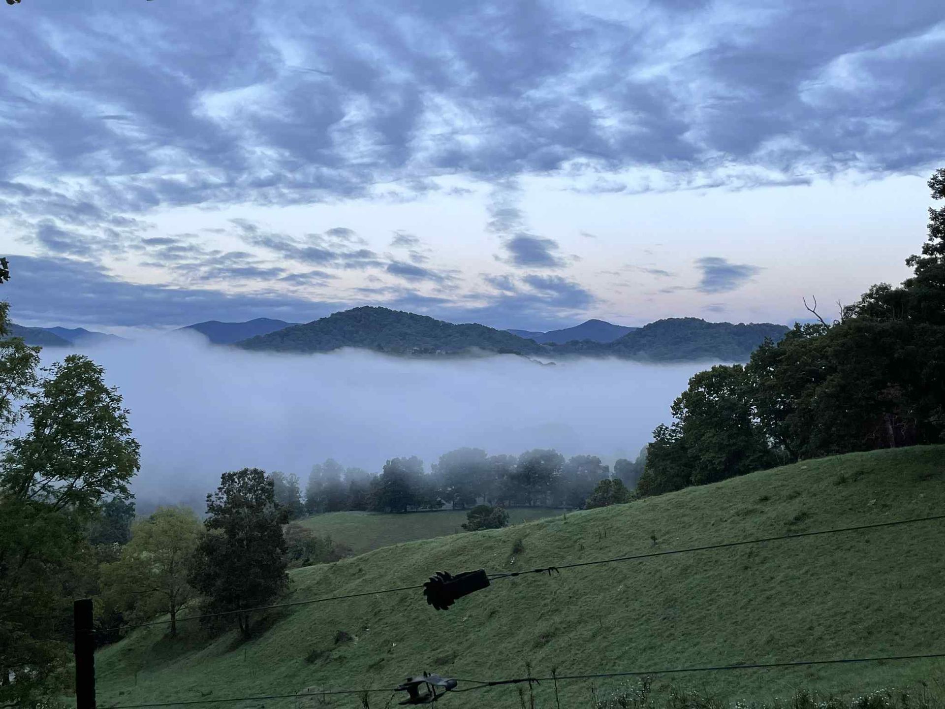 Hilly landscape with fog-filled valley, trees, and cloudy sky at dawn.
